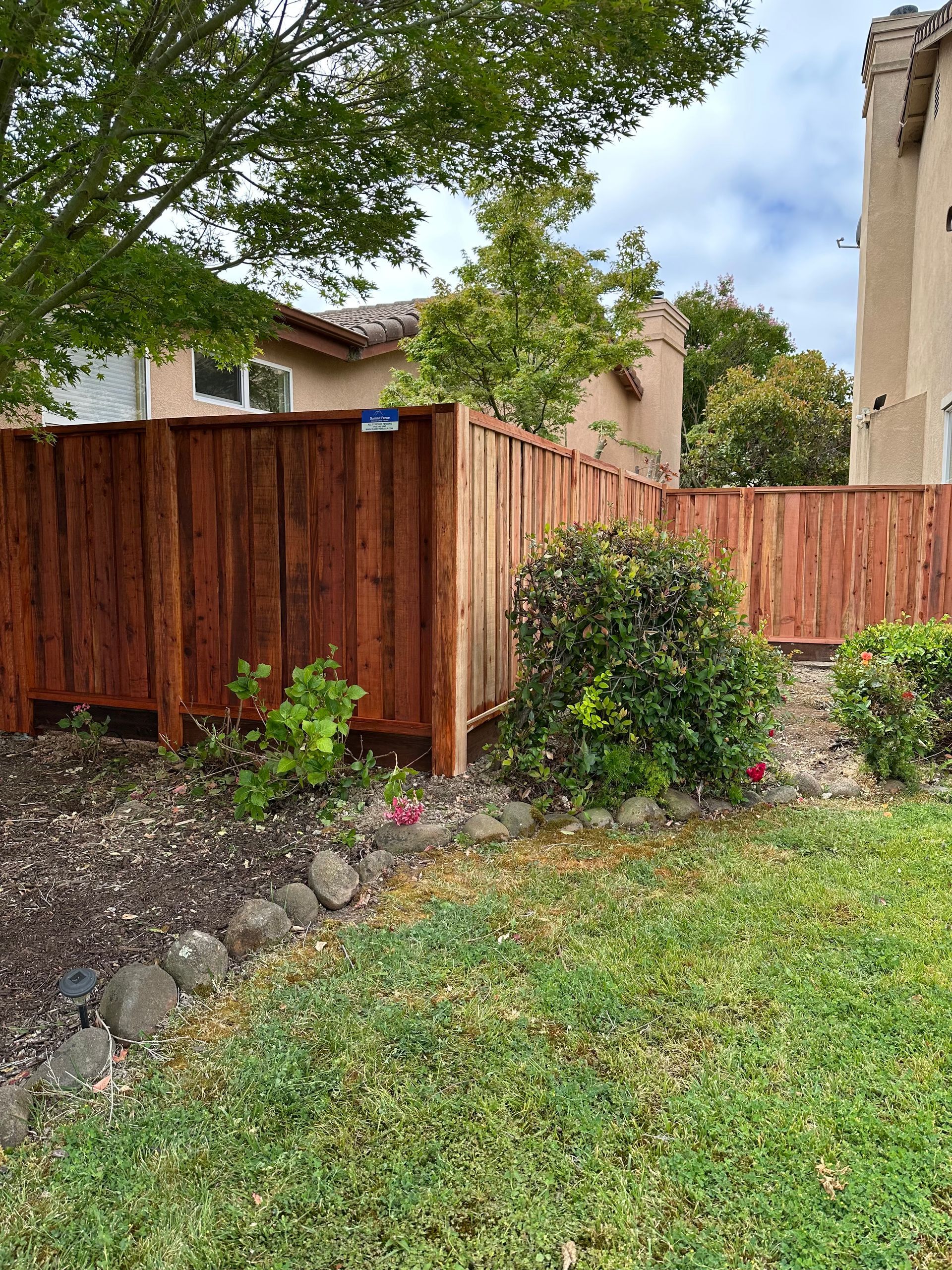 Wooden backyard fence with shrubs, stone border, and green lawn beside a house.