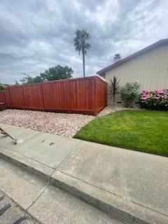Suburban front yard with red wooden fence, gravel strip, green lawn, and a house under cloudy sky