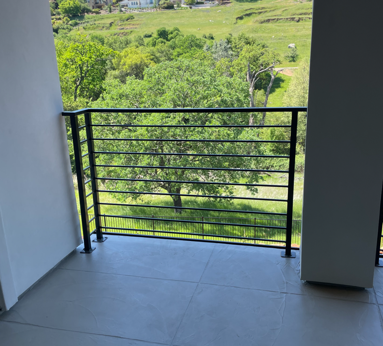 Balcony with black railing overlooking a green landscape with trees and a few buildings.