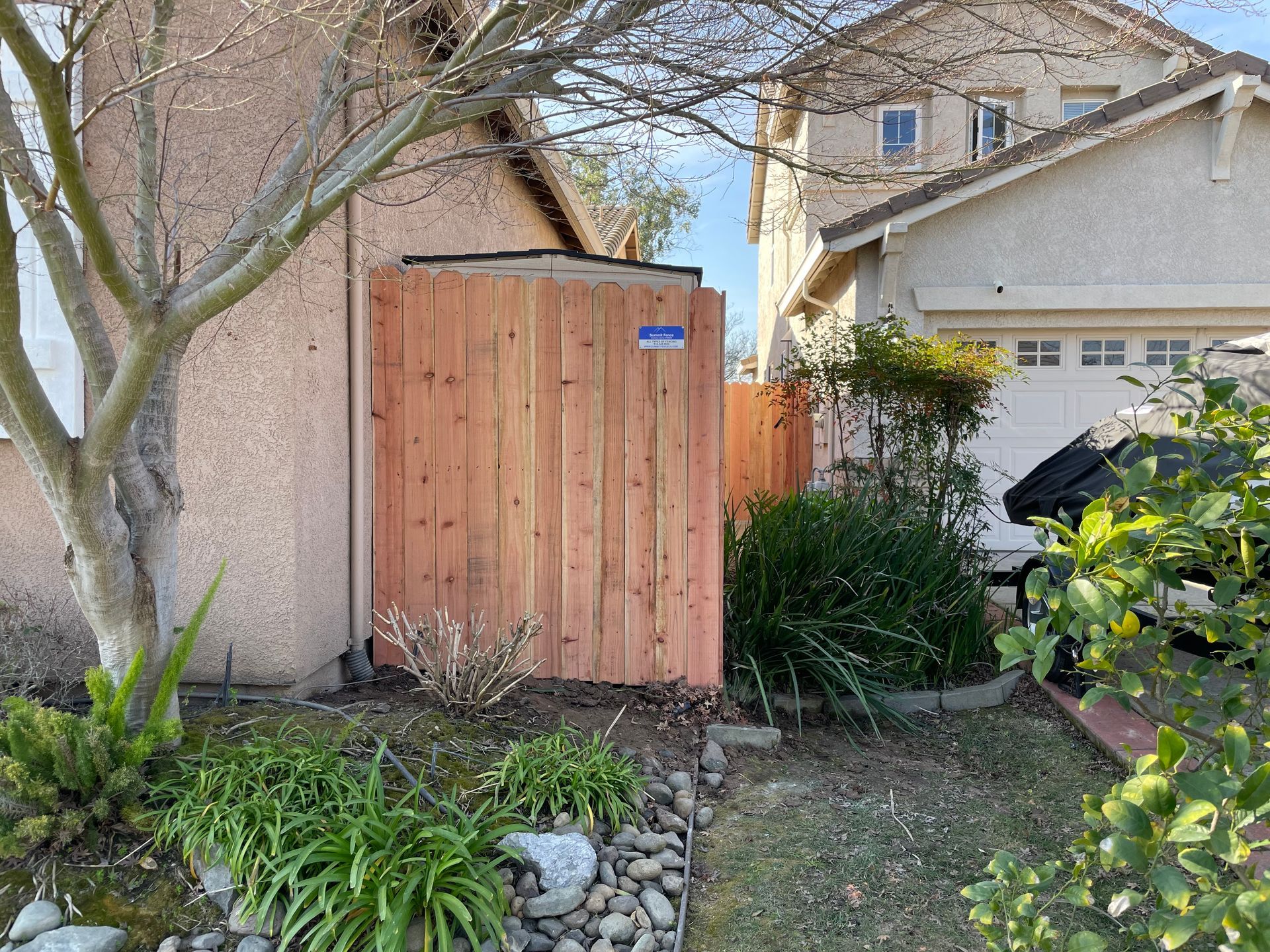 White picket fence surrounds a backyard pool. Plants and rocks border the fence and patio.
