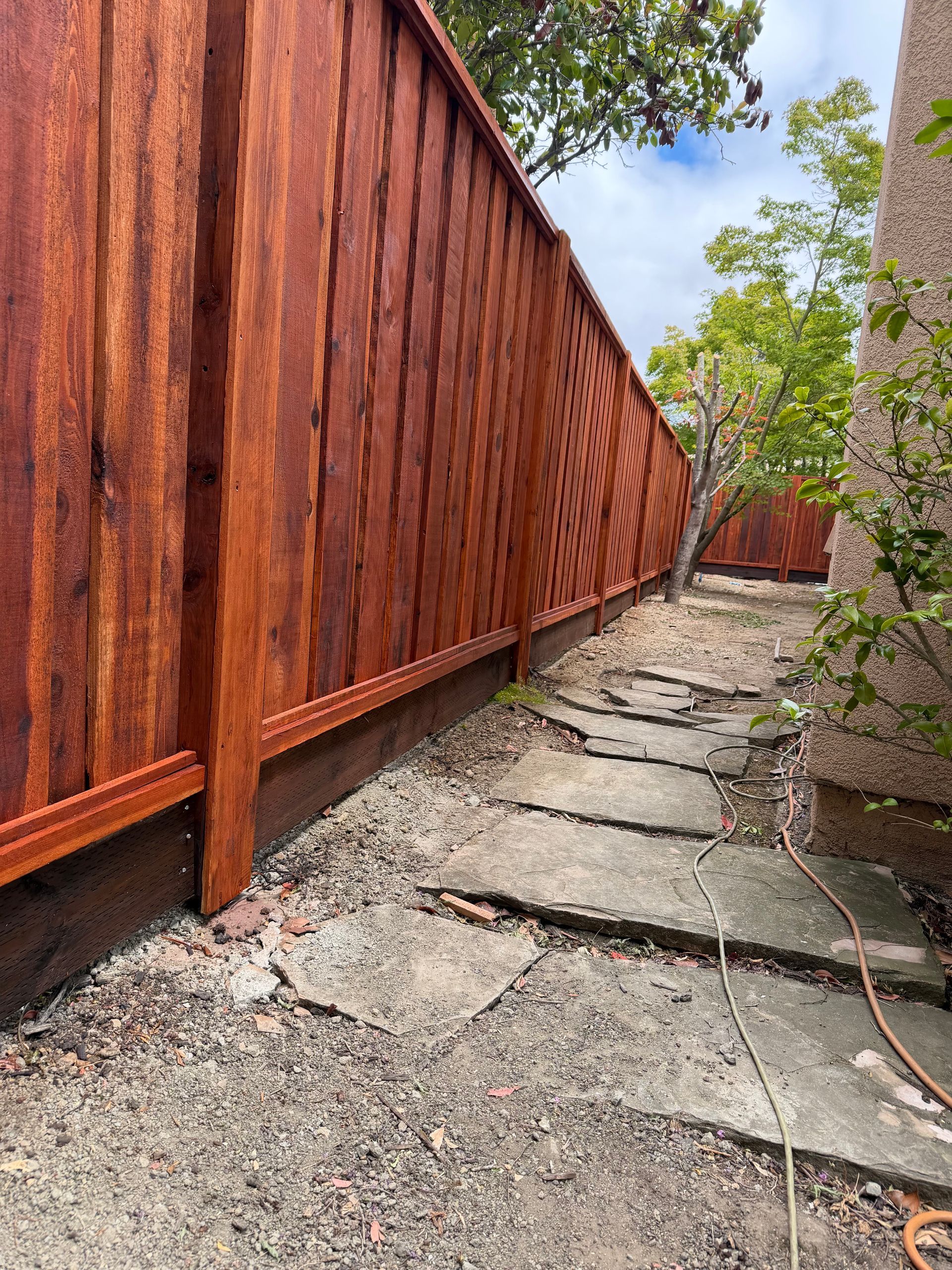 Narrow side yard with red wooden fence, cracked concrete path, and greenery overhead