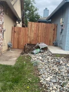 Narrow side yard with a wooden fence, piles of rubble, and a patchy grass path between two houses