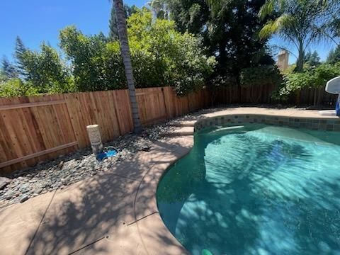 Backyard swimming pool with turquoise water, wooden fence, rocks, and sunlit trees