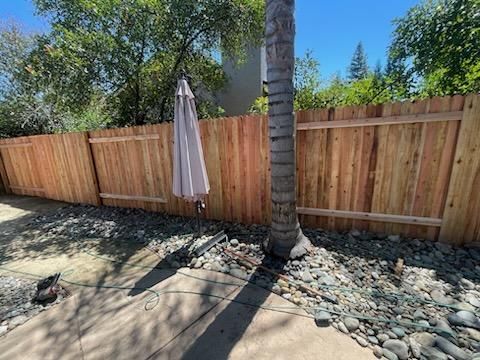 Backyard patio with a closed beige umbrella, palm tree, wood fence, and rock landscaping under bright sun