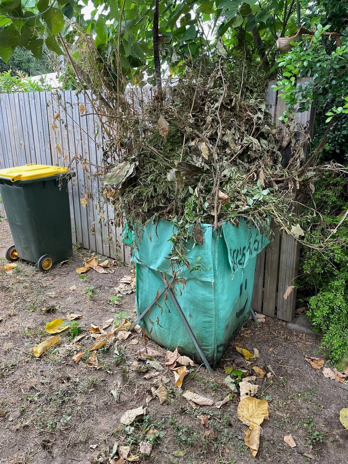 Overfilled Yard Bin with Yard Waste Beside a Fence and Black Trash Bin Outdoors — Green Waste in Thuringowa Central, QLD