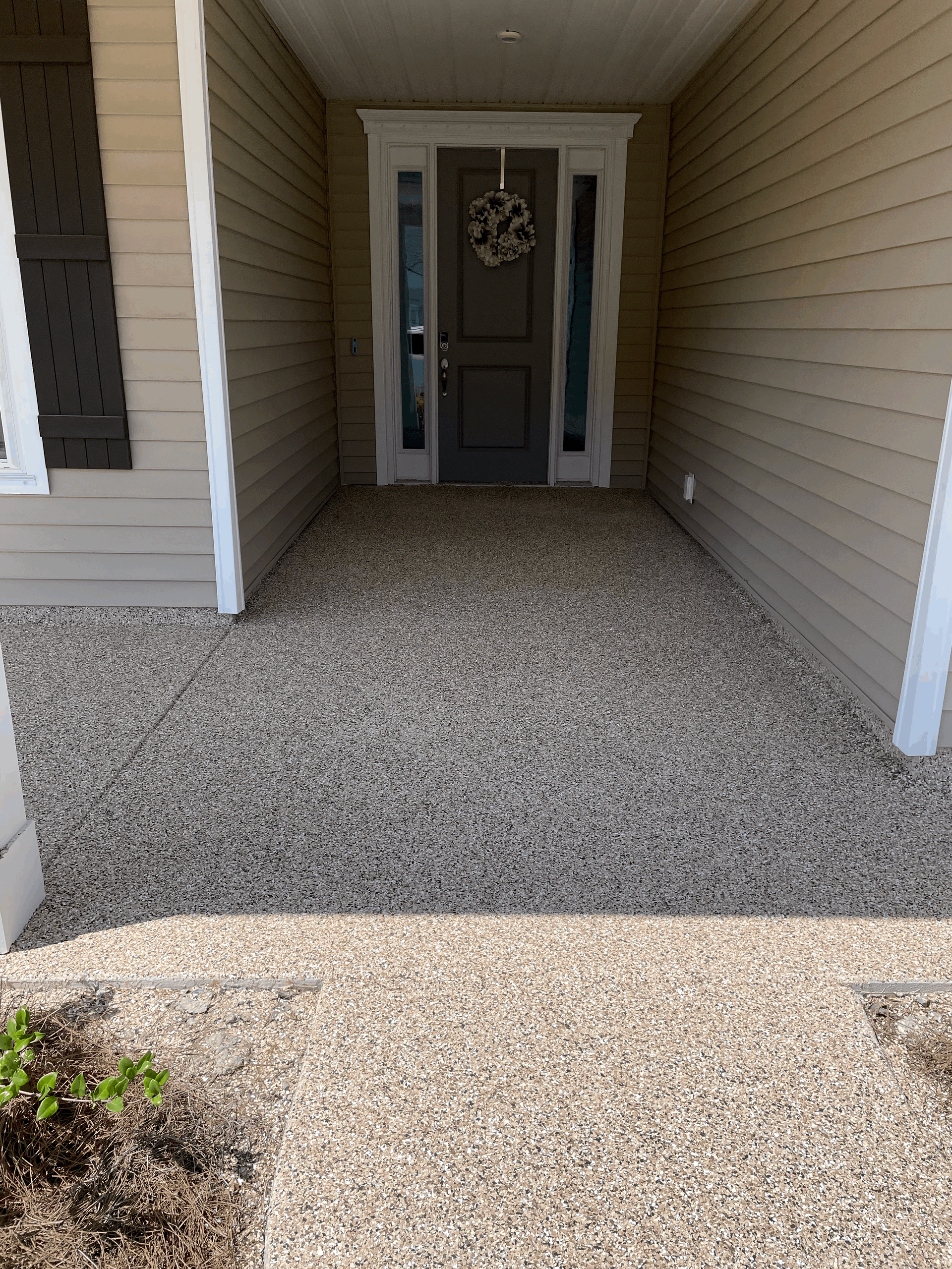 A covered front porch with gravel flooring leading to a dark brown front door with side windows and a circular wreath.