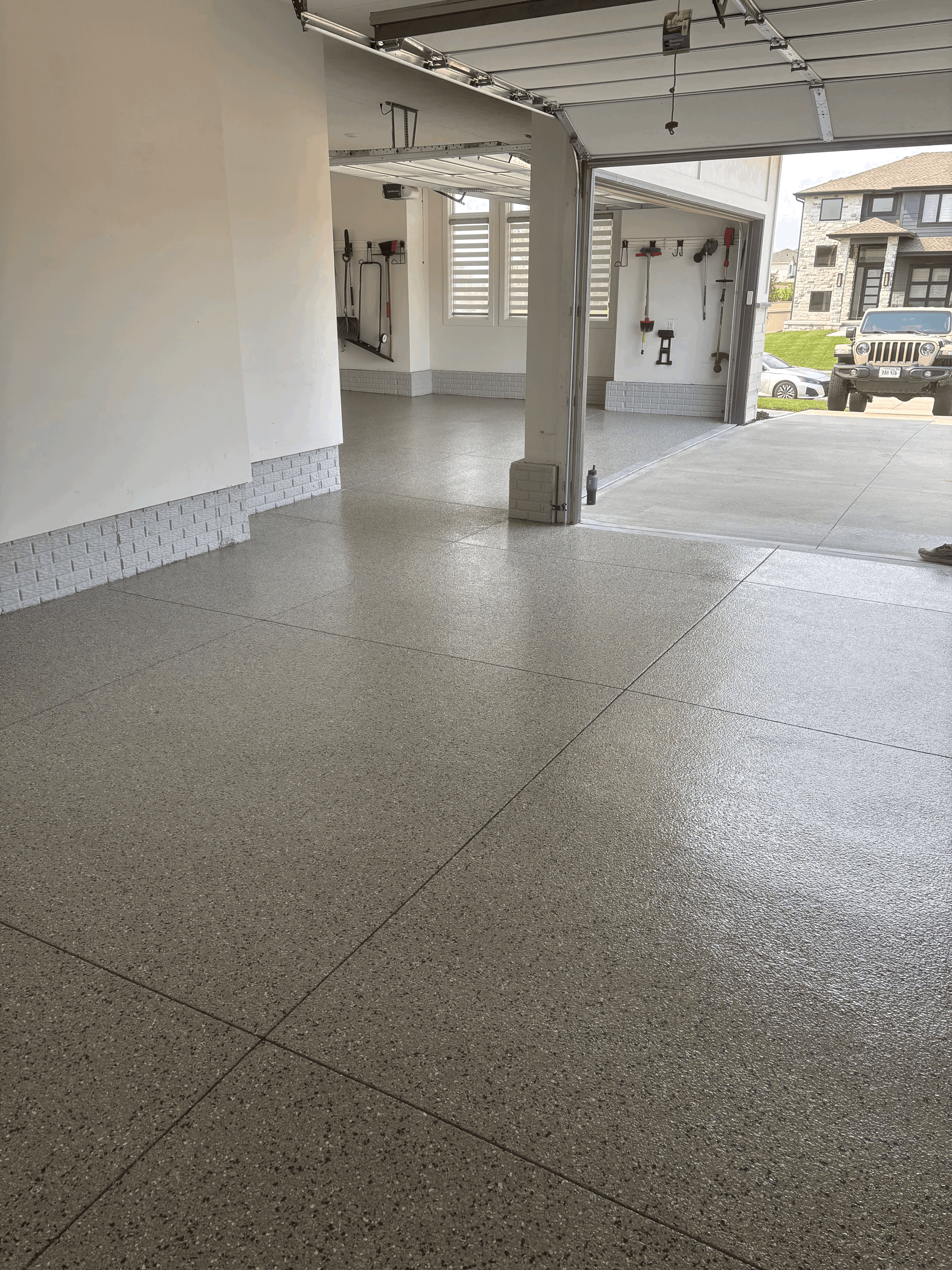 A modern garage interior with light-colored walls, a gray speckled epoxy floor, and a view of a Jeep parked outside.