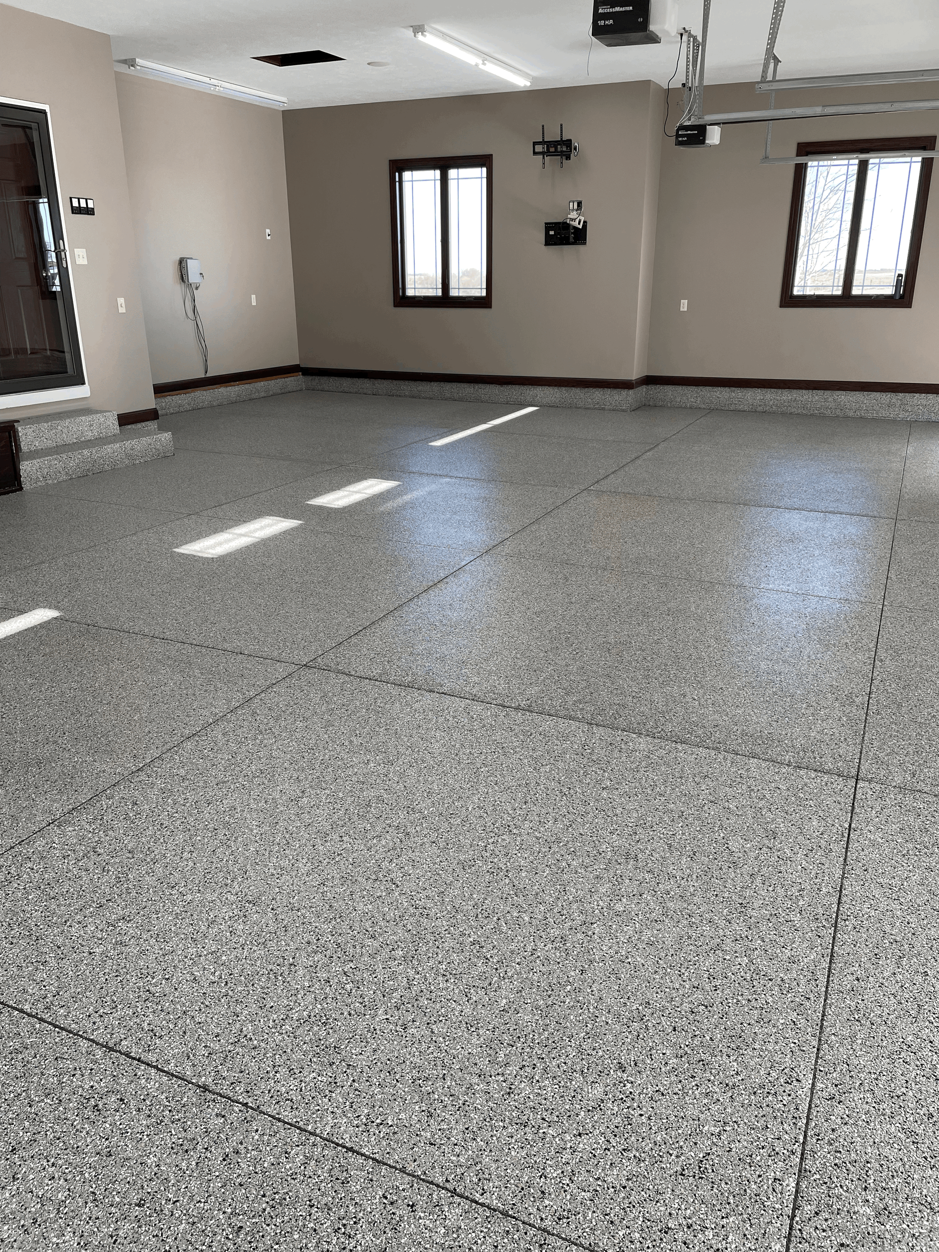 A garage with a polished gray epoxy flake floor, neutral walls, two windows, and overhead lighting fixtures.