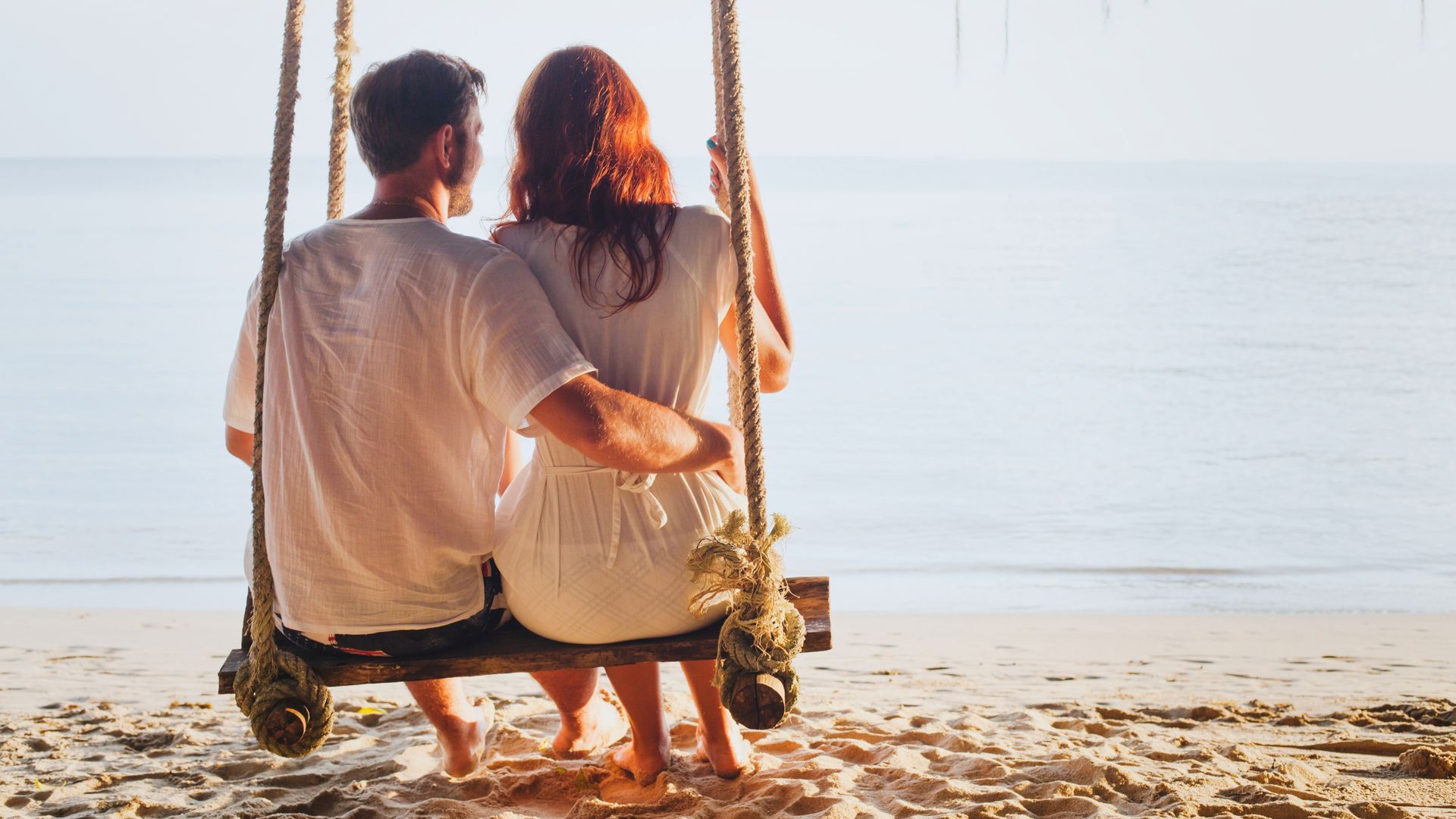 Casal em um balanço, praia, oceano ao fundo. O homem abraça a mulher, ambos vestem branco, luz do sol.