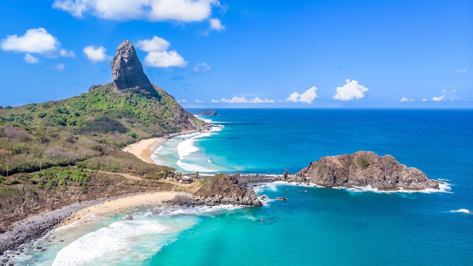 Vista costeira: Praia da Conceição, com água turquesa, costa rochosa e uma exuberante montanha verde sob um céu azul.