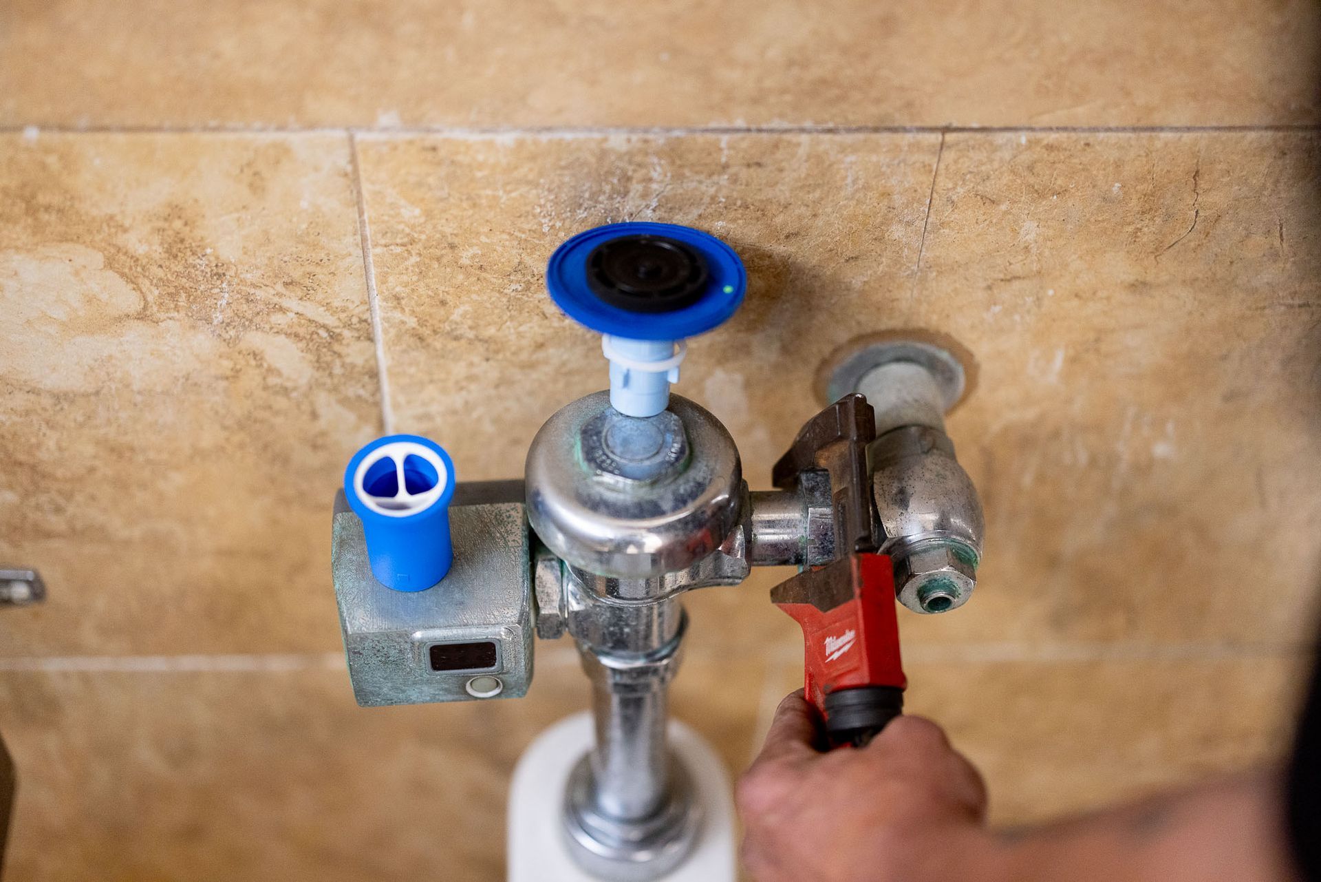 A person using a wrench to repair a silver urinal flush valve against a beige tile wall.