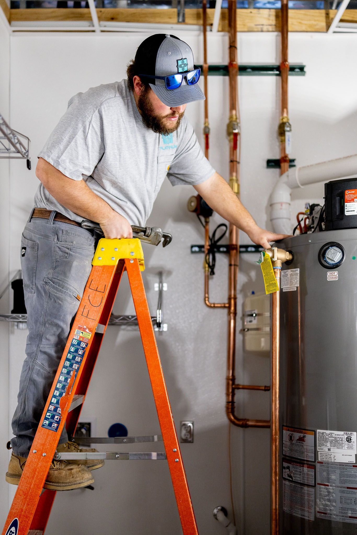 Plumber on a step ladder, working on copper pipes connected to a hot water heater in a utility room.