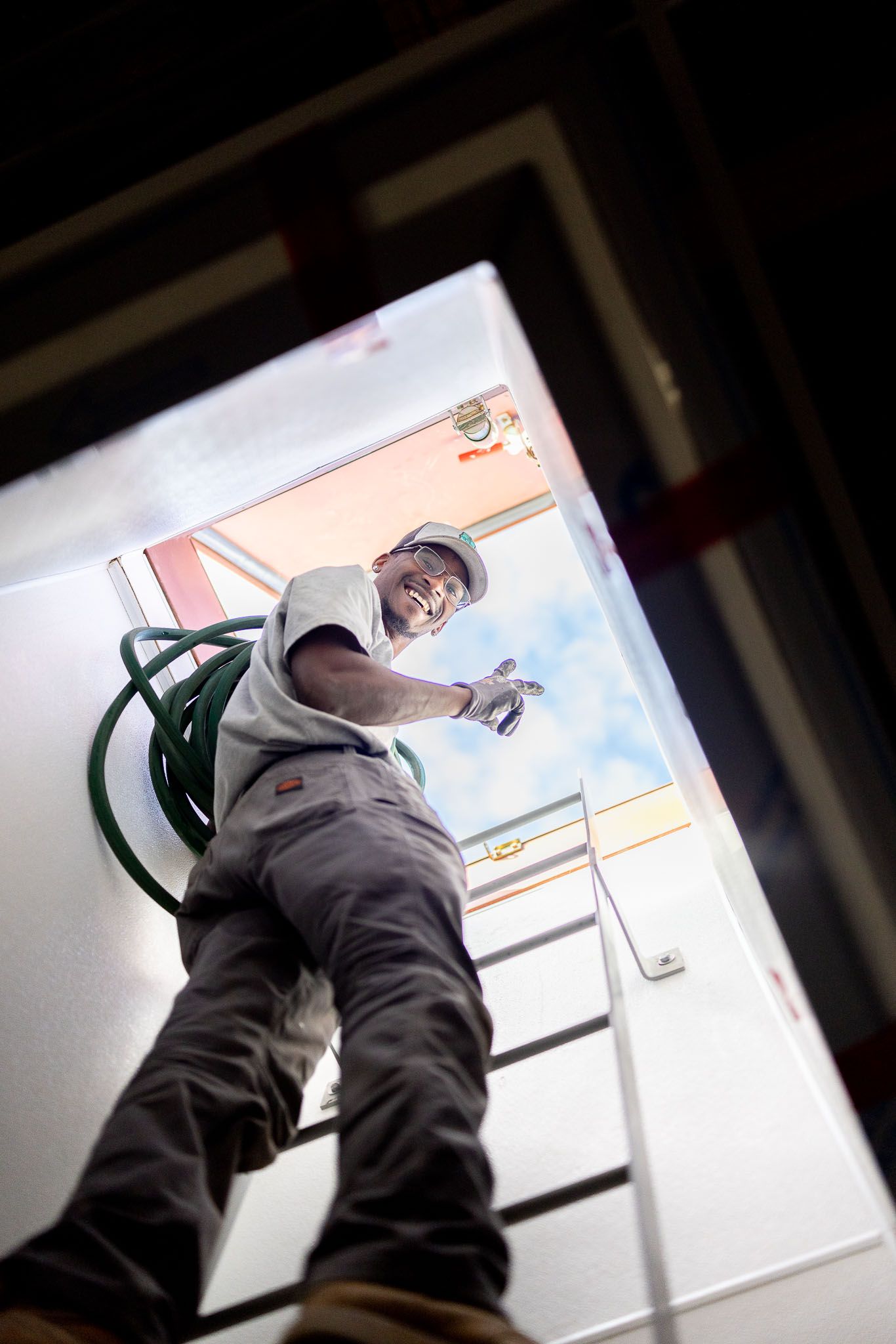 Man climbing a ladder, smiling, reaching toward an open skylight, green hose attached.