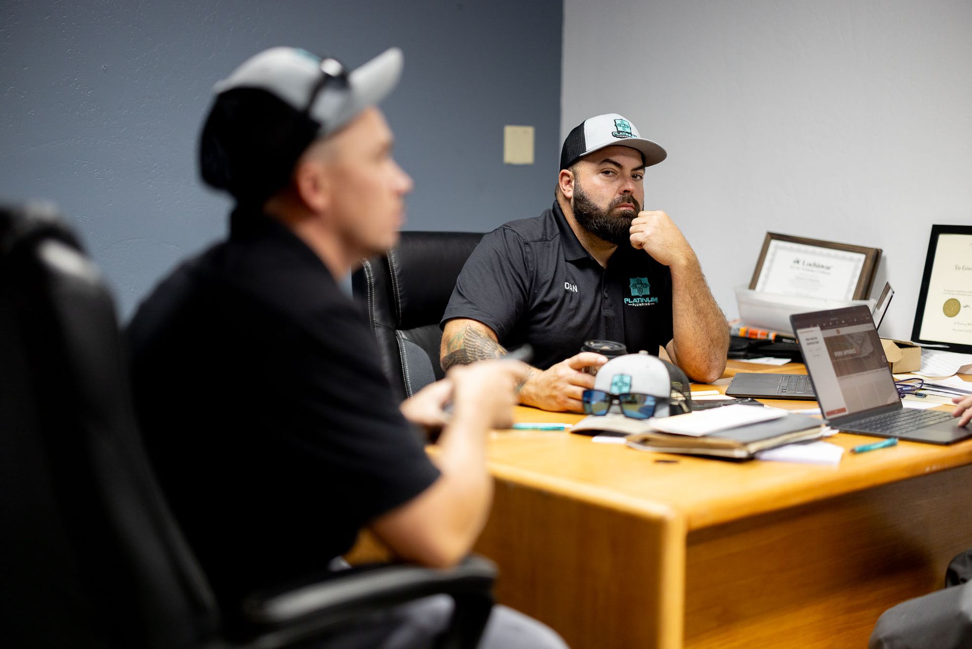 Two men are sitting at a desk in an office talking to each other.