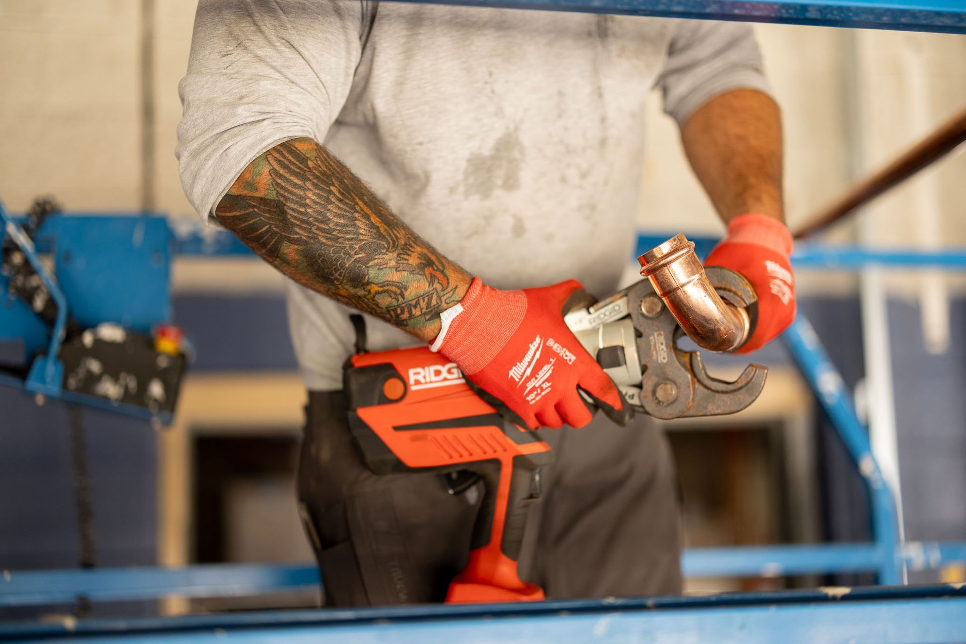 Plumber in red gloves using a copper pipe cutter. Gray shirt, visible arm tattoo, with red power tool at waist.