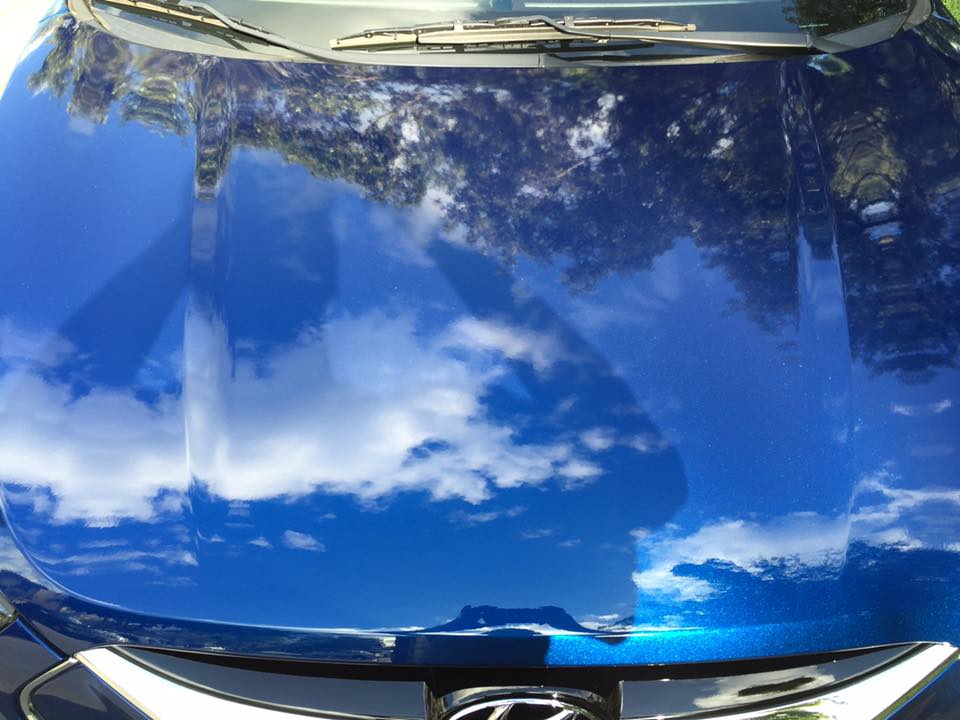 The hood of a blue car is reflecting the sky and clouds
