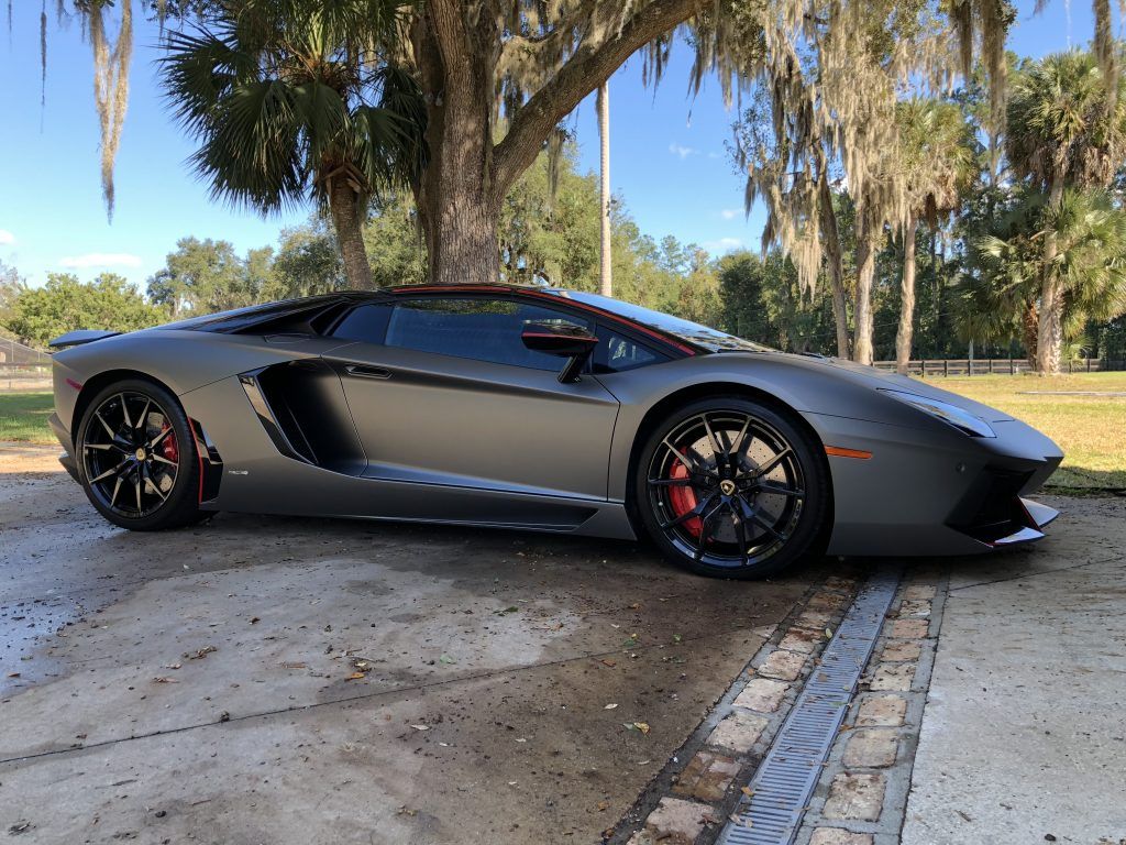 A gray lamborghini aventador is parked in a driveway next to a tree.
