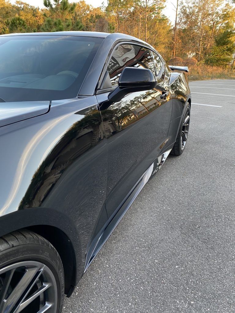 A black mustang is parked in a parking lot with trees in the background.