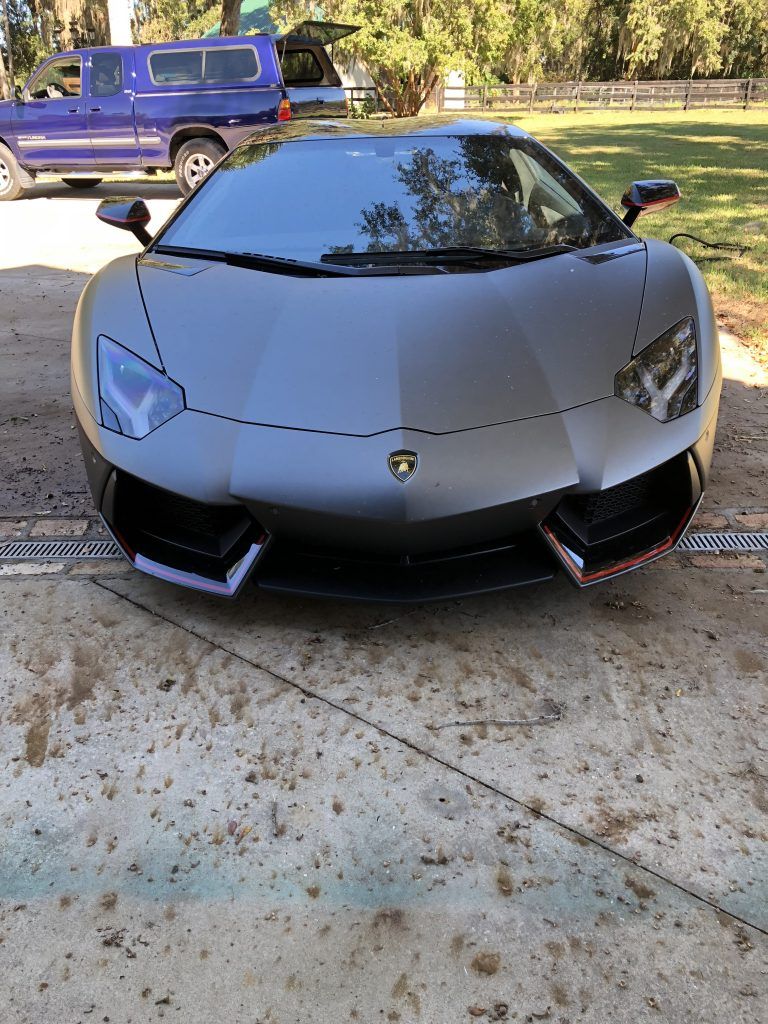 A gray lamborghini aventador is parked in a driveway next to a blue truck.