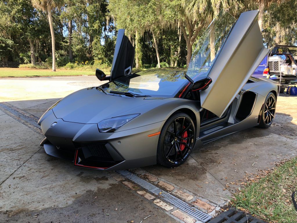 A gray lamborghini aventador is parked on the side of the road with its doors open.