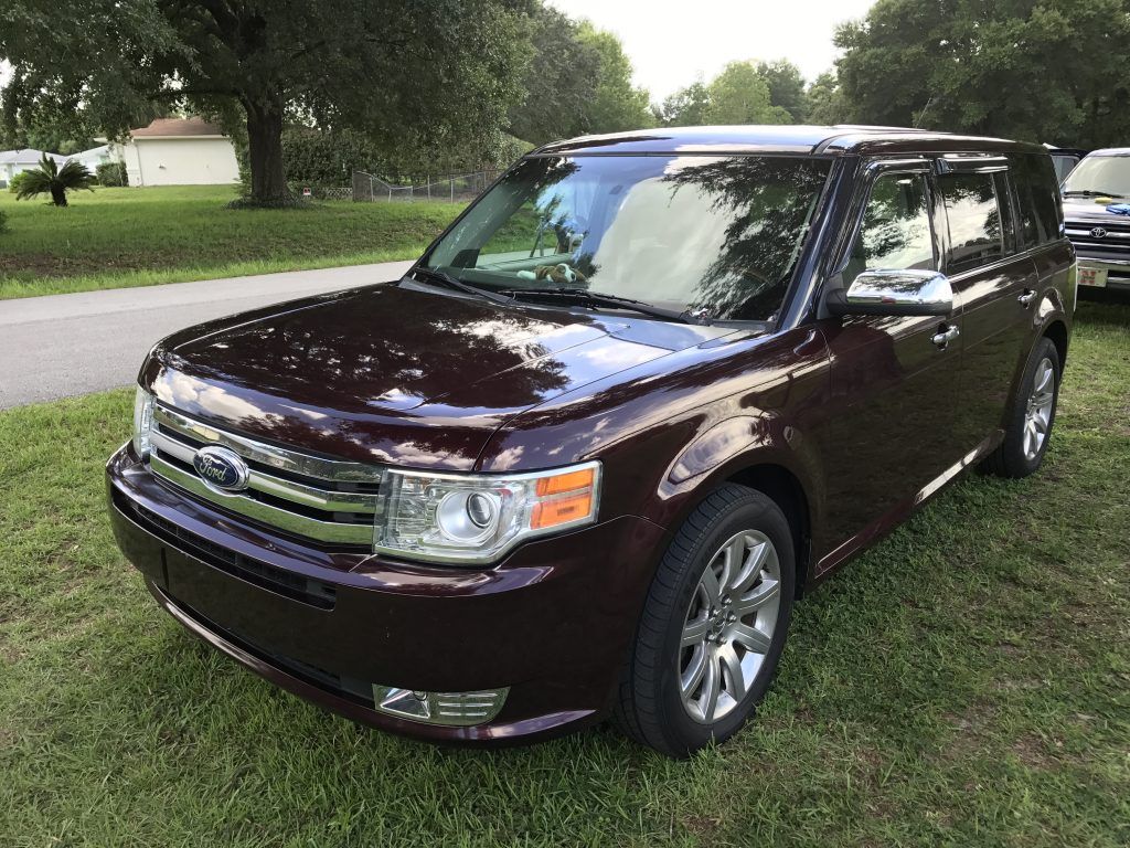 A burgundy ford flex is parked in the grass on the side of the road.