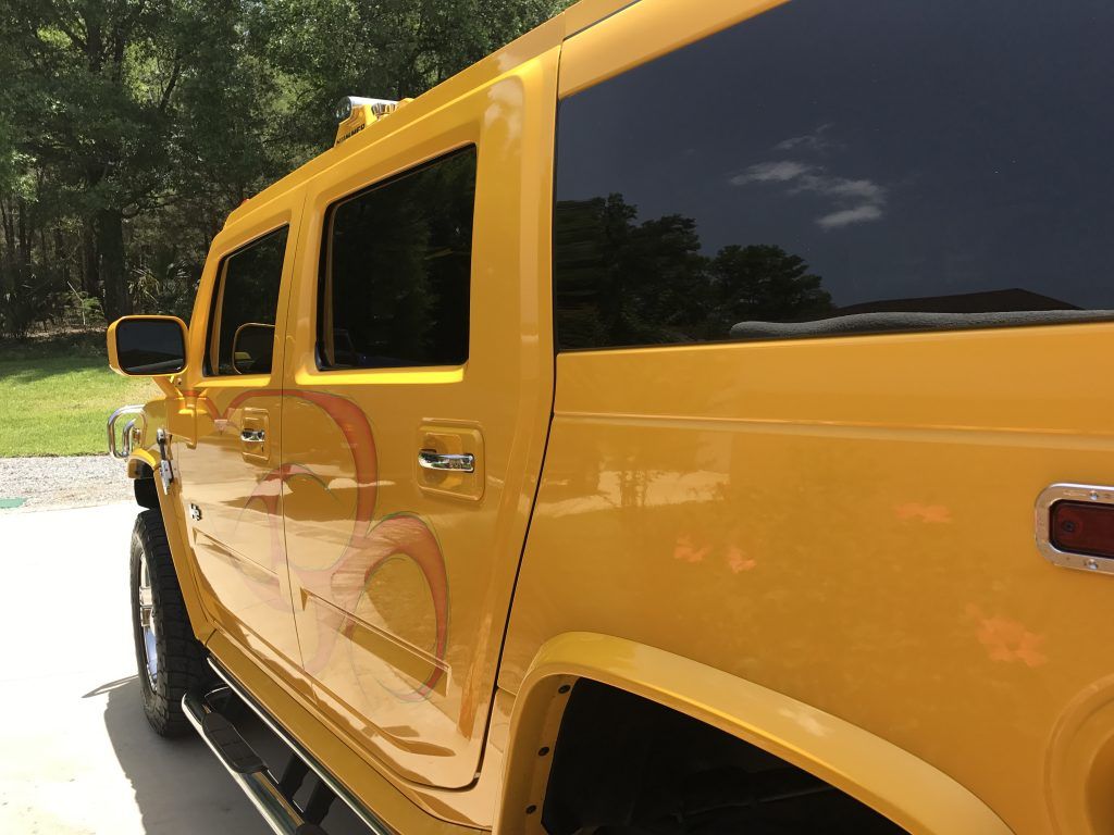 A yellow hummer is parked in a driveway with trees in the background