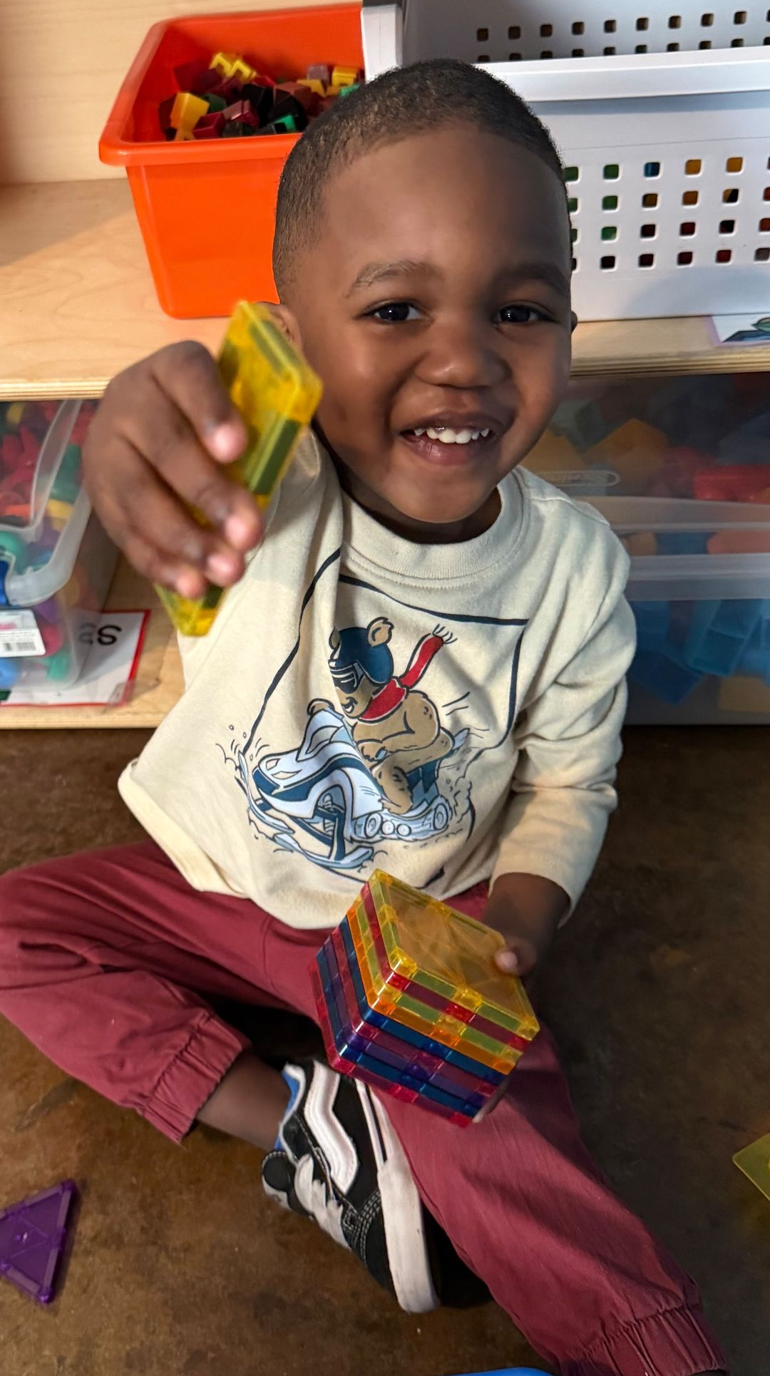 Young child smiles, holding colorful geometric shapes while sitting on a floor.