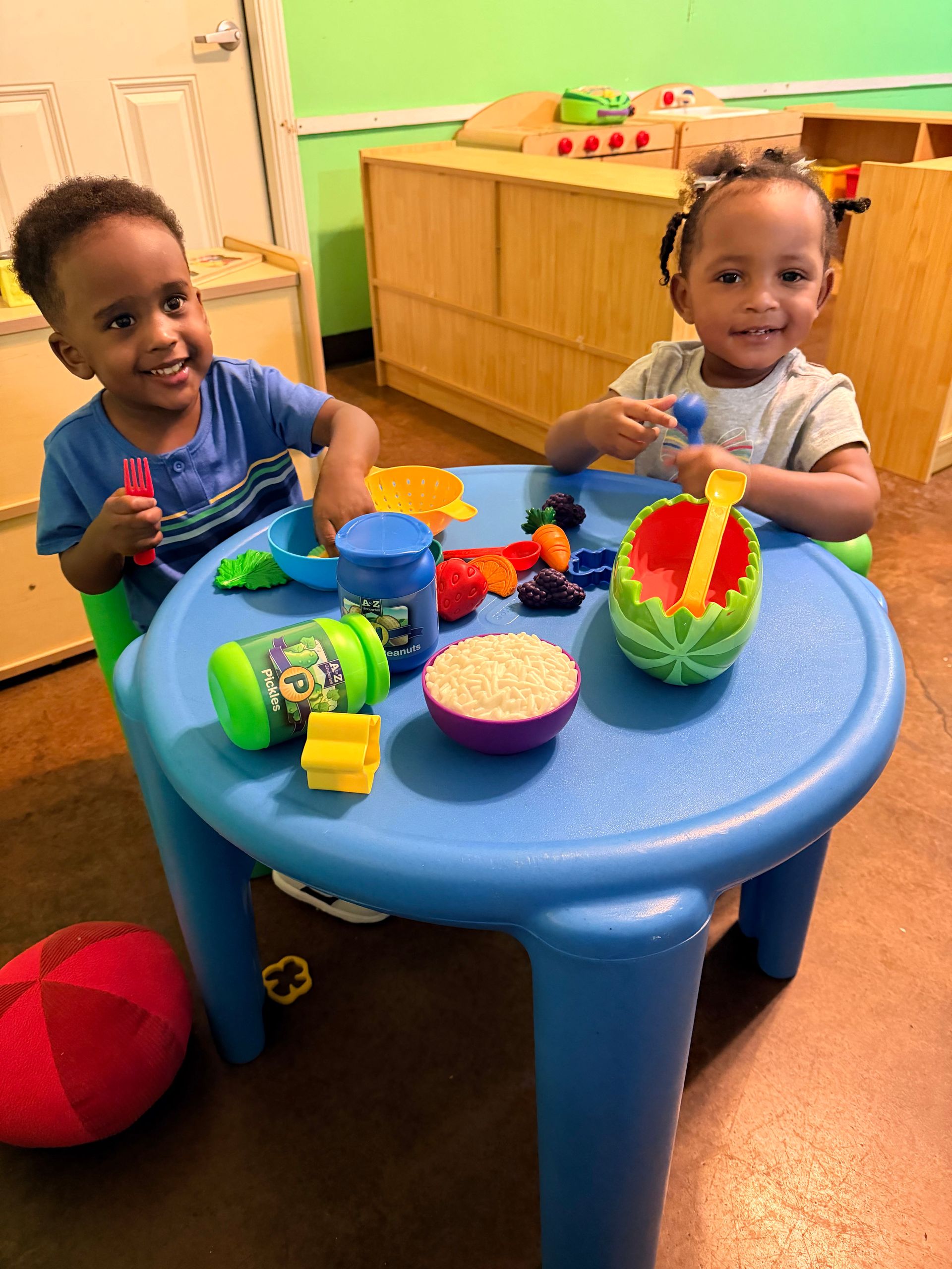 Two children at a blue table, playing with toys. Smiling in a brightly lit room.