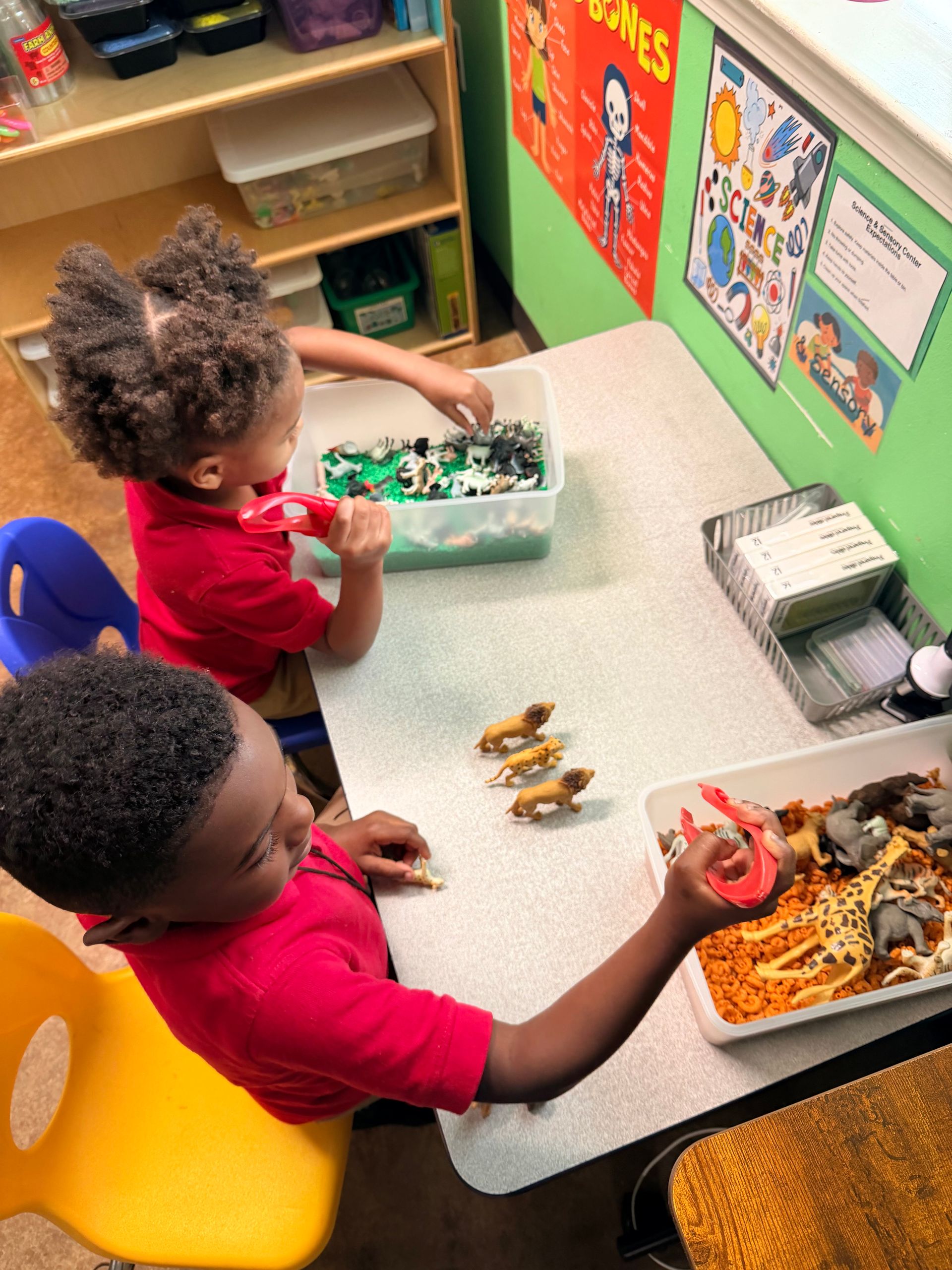 Two children at a table playing with toy animals and building blocks in a classroom.
