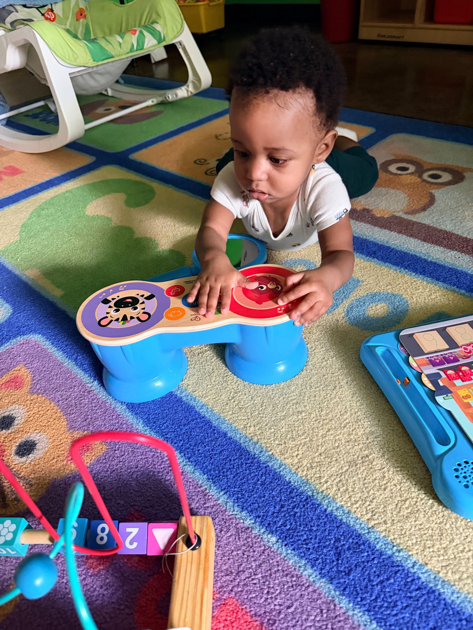 Baby playing with toys on a colorful rug in a room, reaching for a blue and red toy.