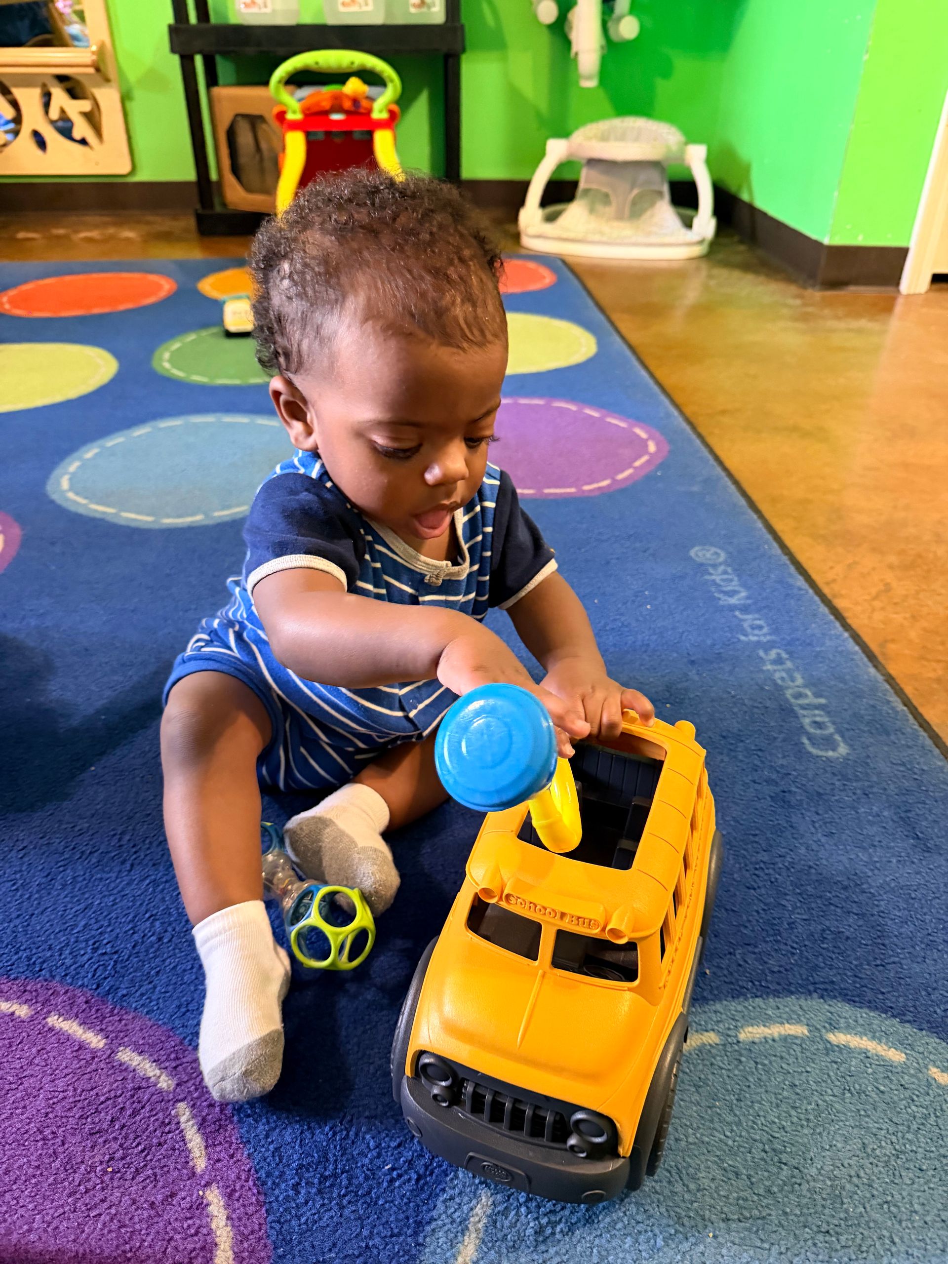 Child playing with yellow toy school bus on colorful rug.