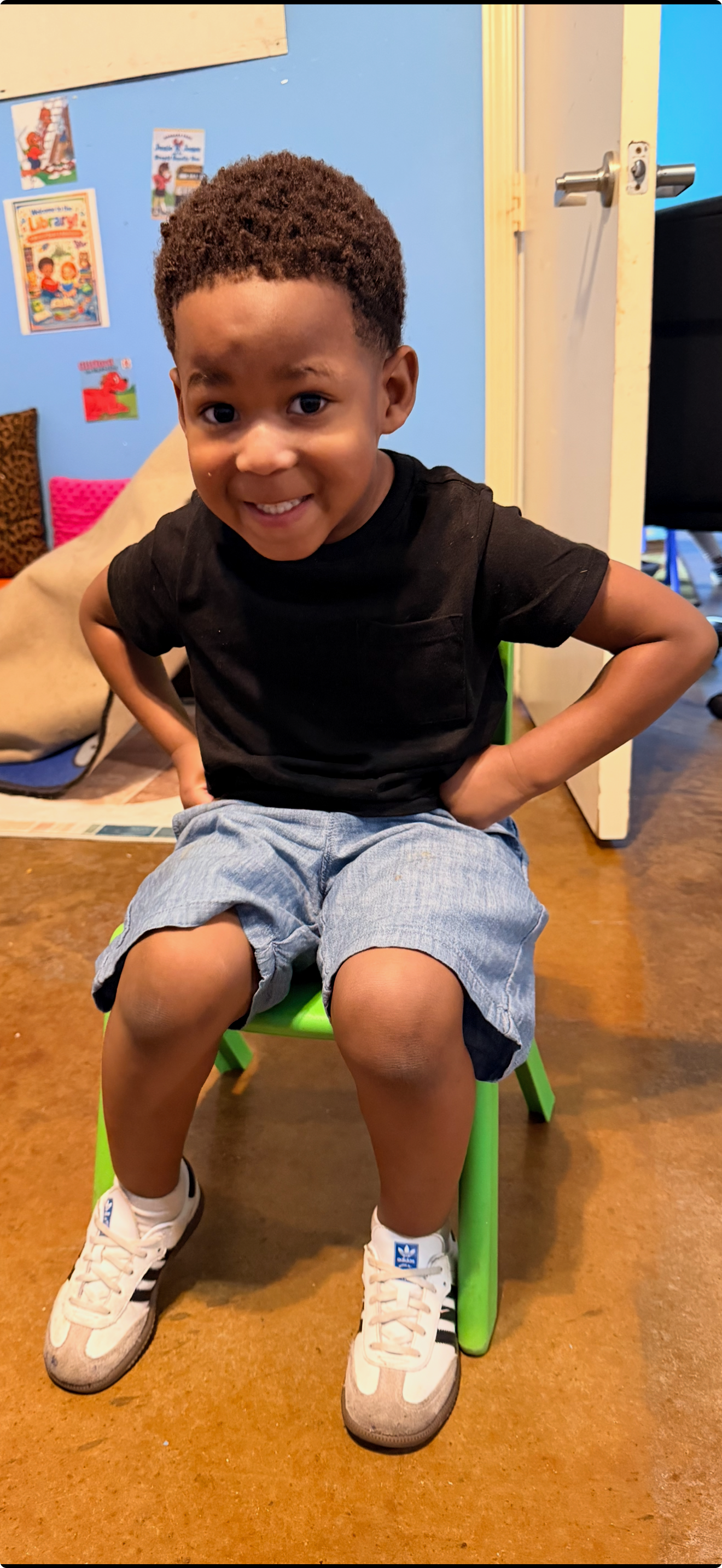 A young child with hands on hips, smiling, wearing a black shirt, shorts, and sneakers, sitting on a green chair.