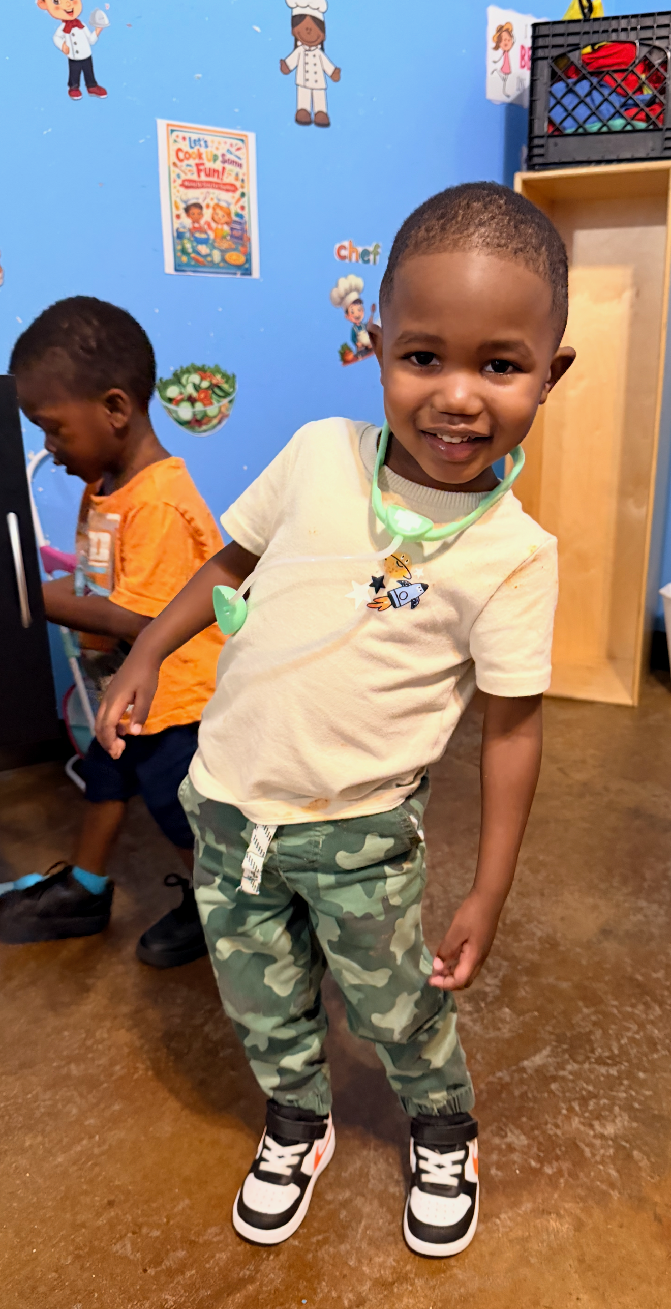 Smiling child in camouflage pants and cream shirt with another child in the background.