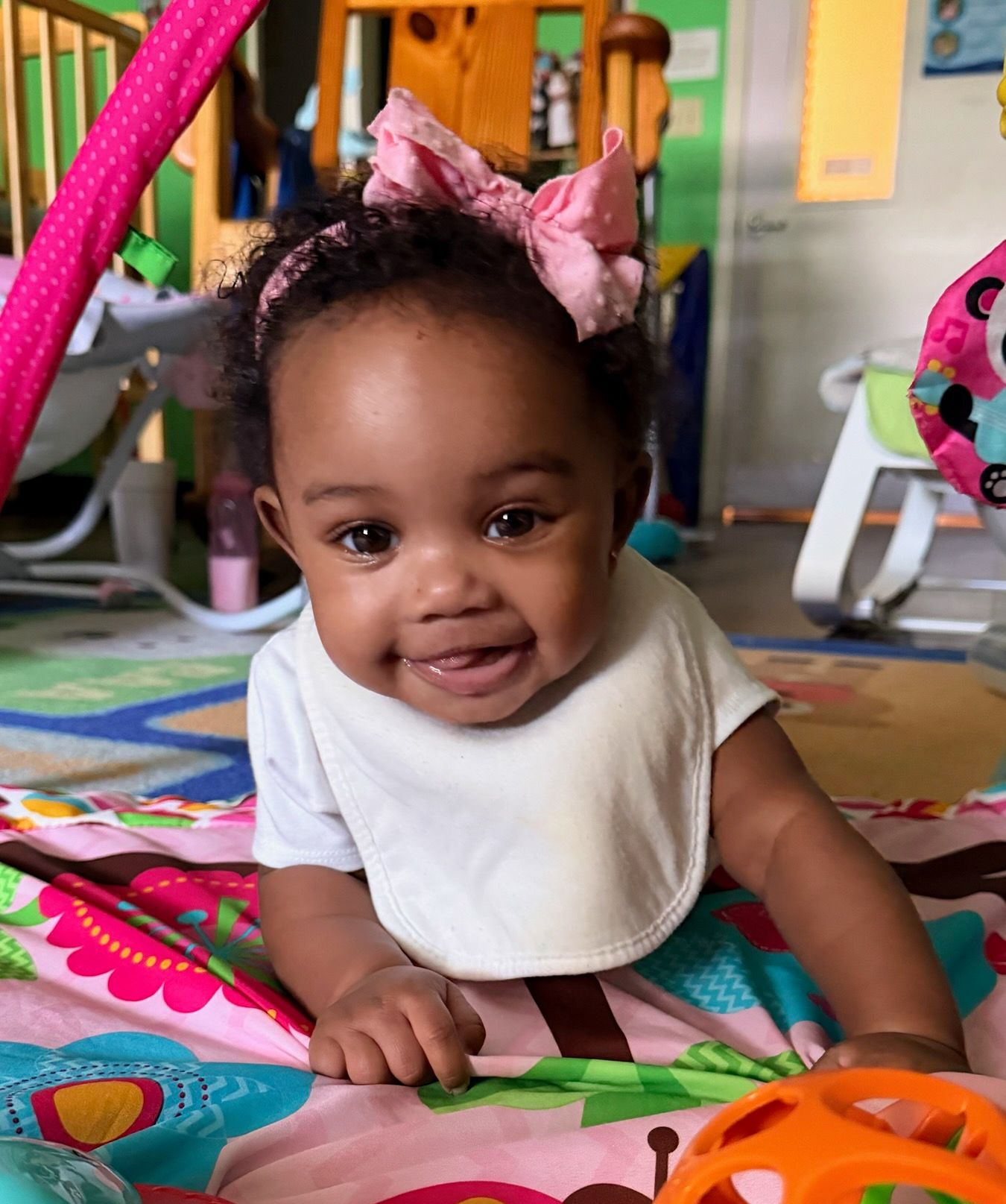 Baby on colorful playmat, wearing bib and pink bow. Smiling.