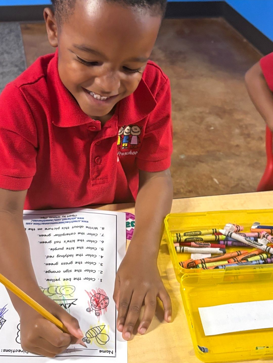 Smiling child in red shirt coloring at a desk, crayons in a yellow container.