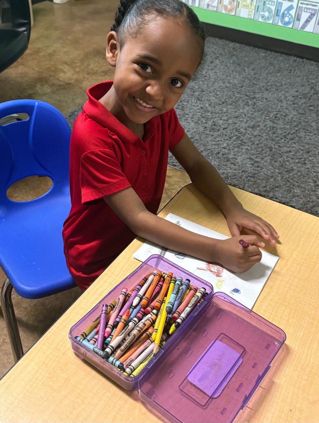 Girl in red shirt smiles while coloring at a desk with crayons. 