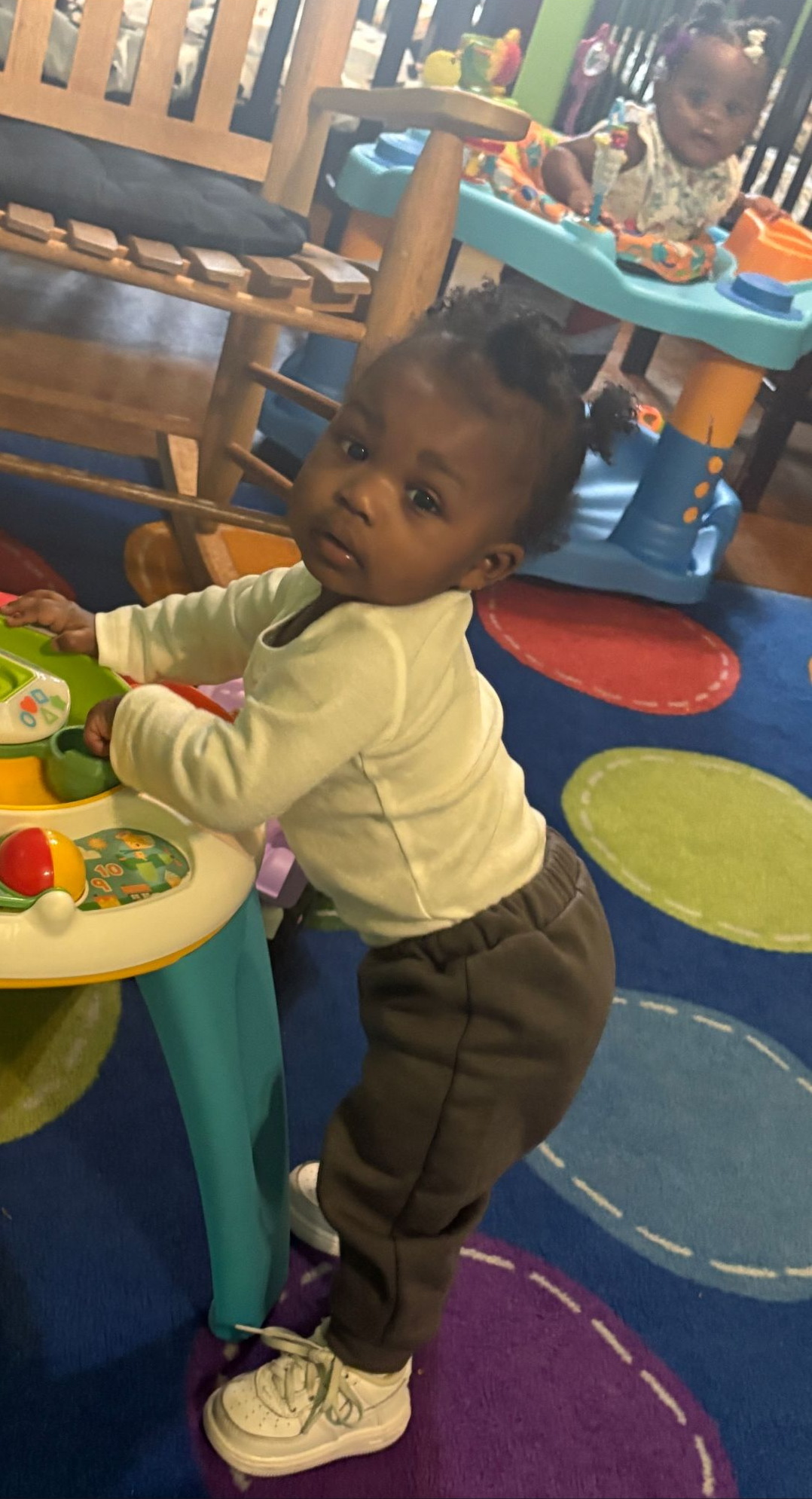 A child stands, holding a toy table, looking towards the camera. Indoors, multi-colored rug.