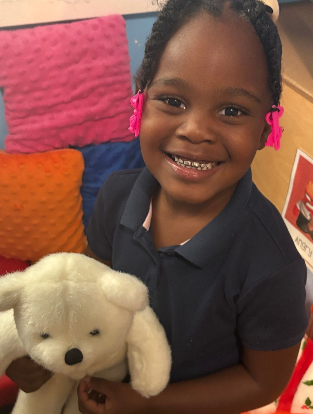 Smiling child holding a white stuffed animal; pink hair clips, blue shirt, colorful pillows in the background.