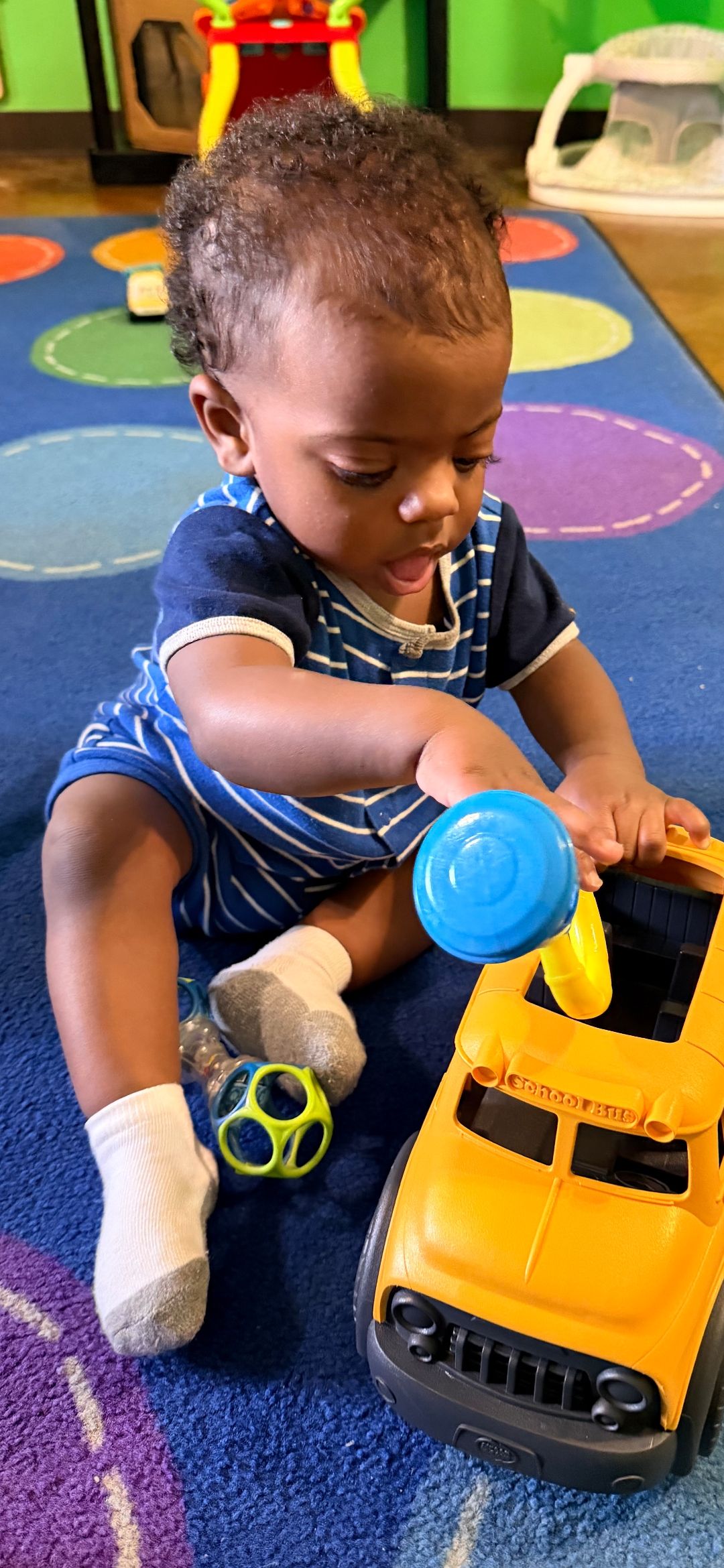 A baby sitting on a colorful mat playing with toys, including a toy truck and blue rattle.