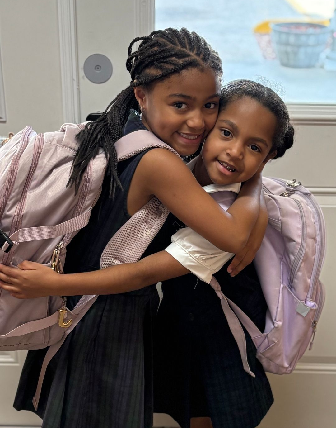 Two girls in school uniforms hug, wearing backpacks.