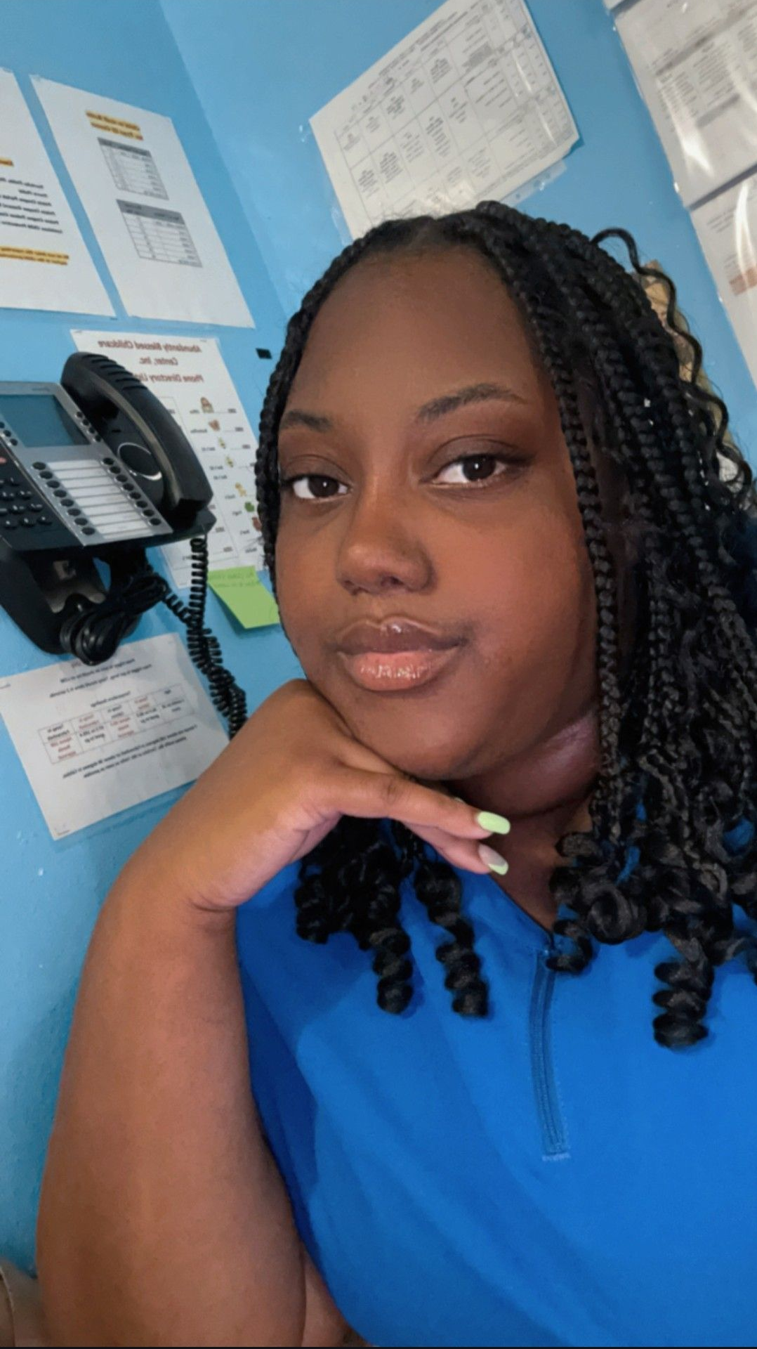 Woman with braided hair, rests hand on chin, smiling. Blue scrubs and a phone in background.
