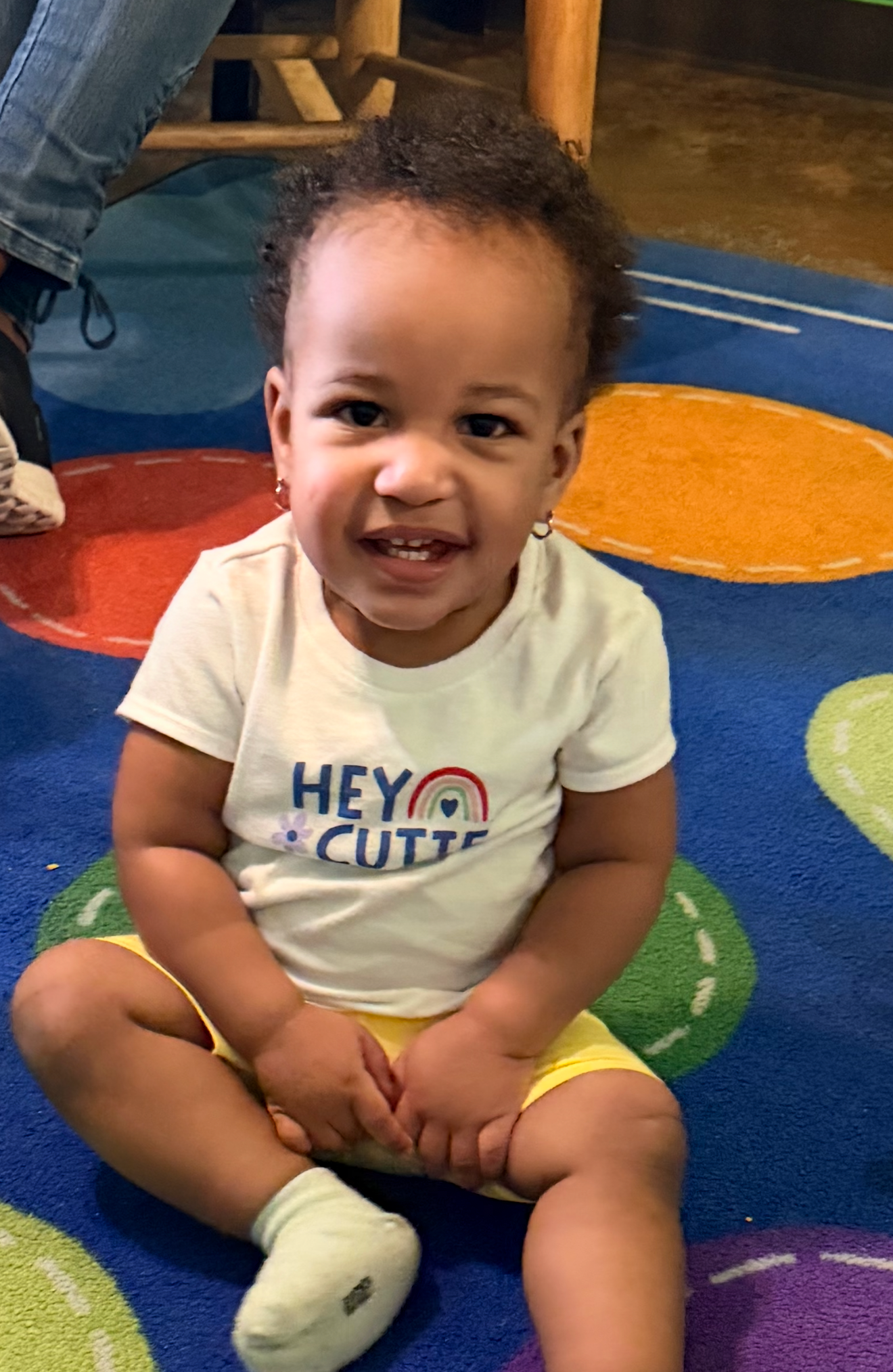 Child sitting on a colorful rug, wearing a white shirt and yellow shorts, smiling.