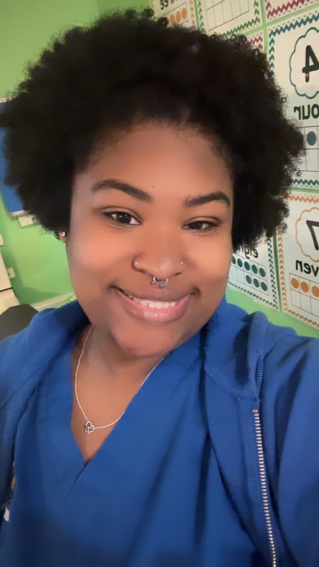 Woman with dark curly hair smiles, wearing blue scrubs, indoors, green wall backdrop.