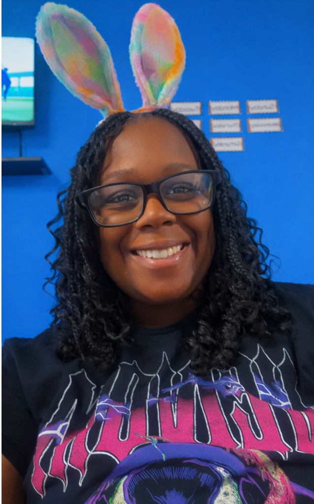 Person wearing bunny ears smiles in front of a blue wall, wearing glasses and a graphic t-shirt.