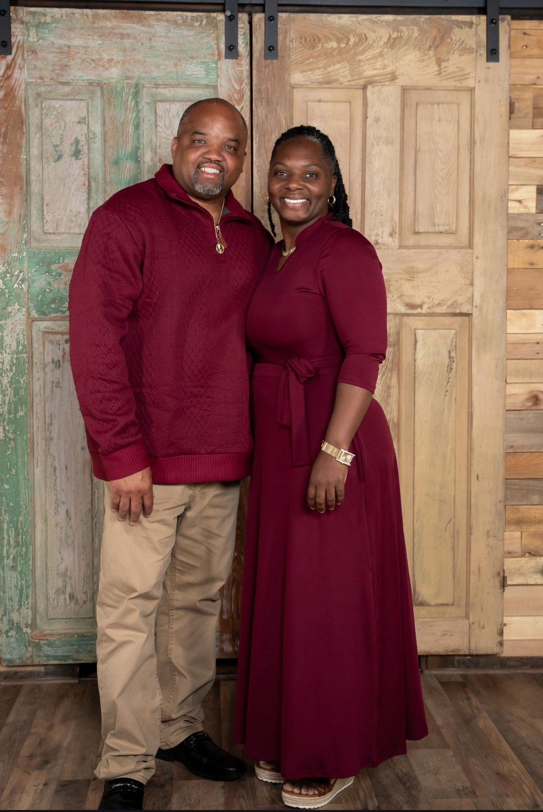 Couple in maroon outfits smiling, standing in front of rustic wooden doors.