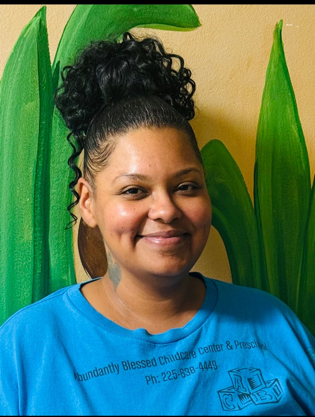 Woman with dark curly hair in a bun smiling, wearing a blue shirt. Background features green leaves.