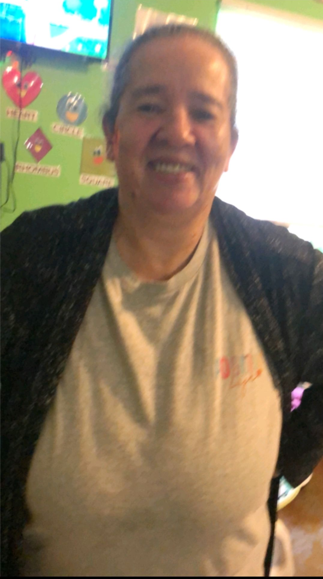Woman wearing gray shirt and dark sweater smiles in front of green wall with colorful shapes.