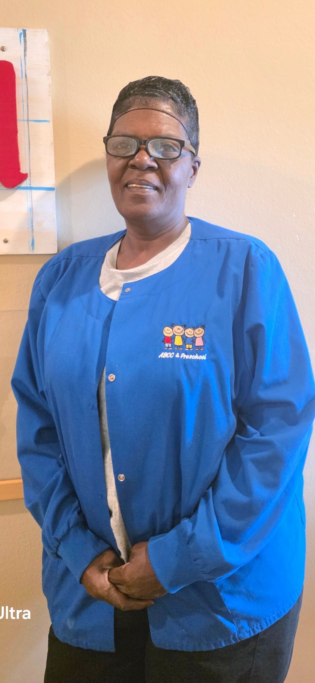 Person wearing glasses and a blue jacket smiles. They stand inside a room with a beige wall.