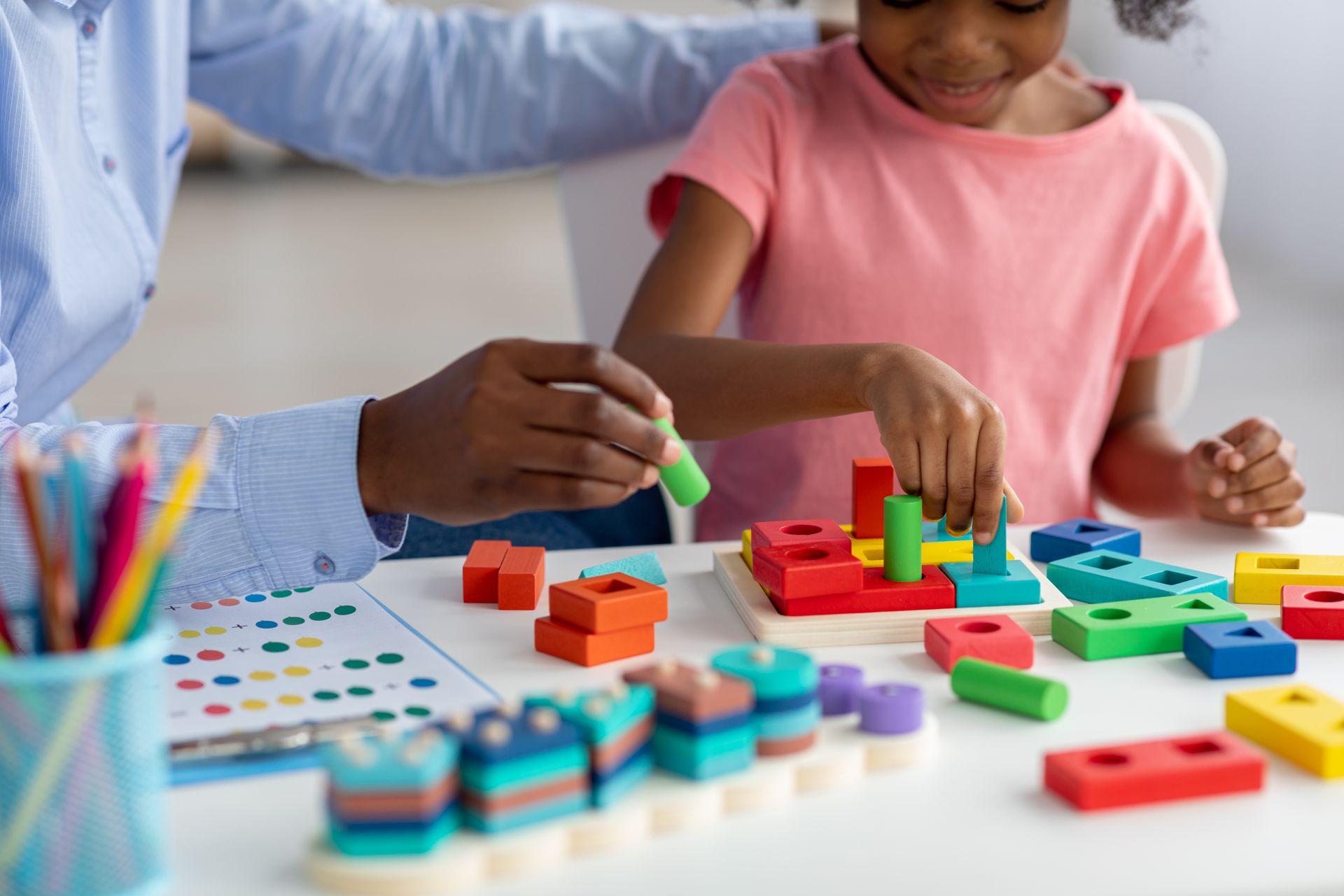 Child and adult playing with colorful shape-sorting blocks at a table.