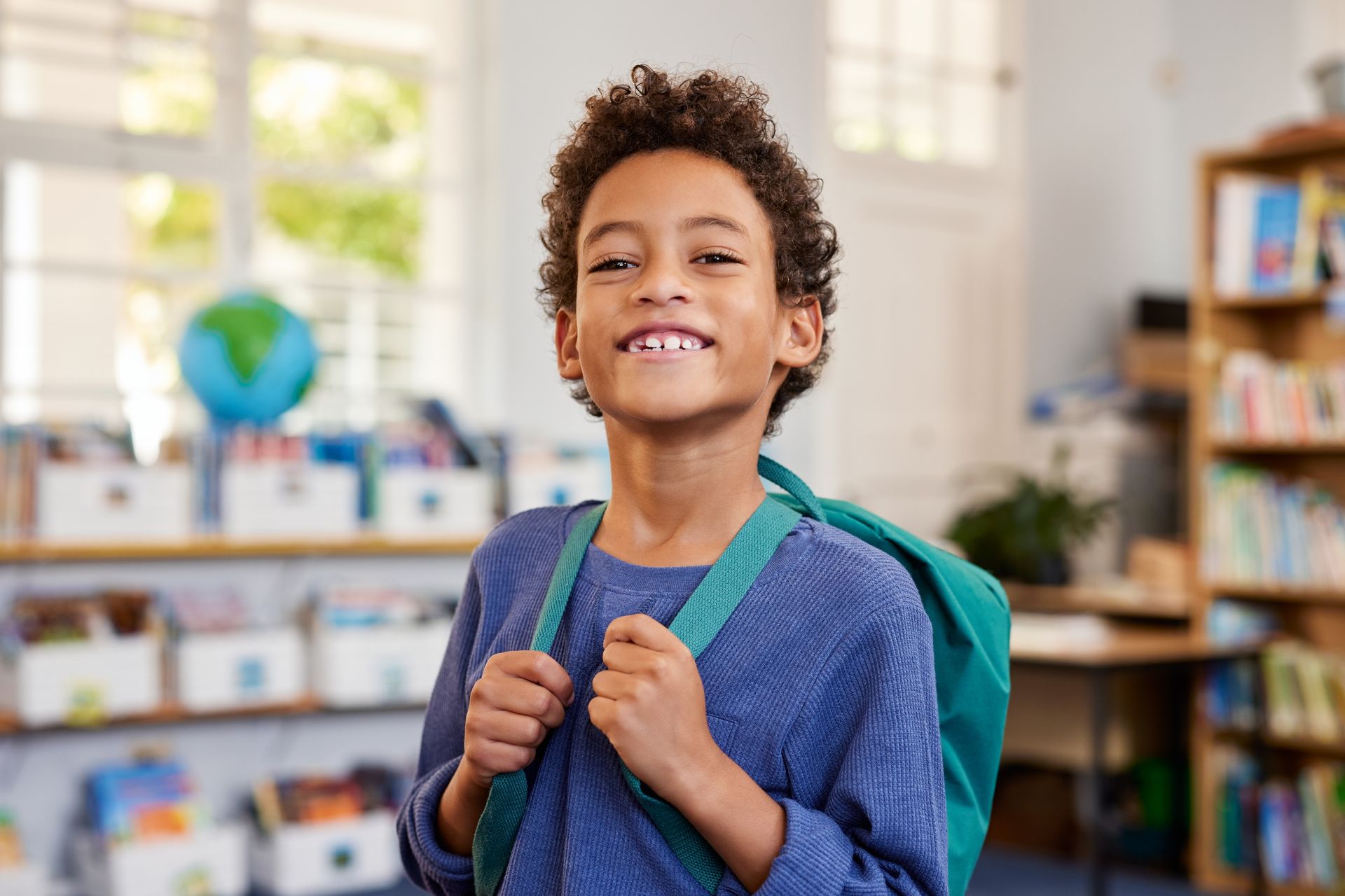 Smiling child in a blue shirt with a backpack in a classroom.
